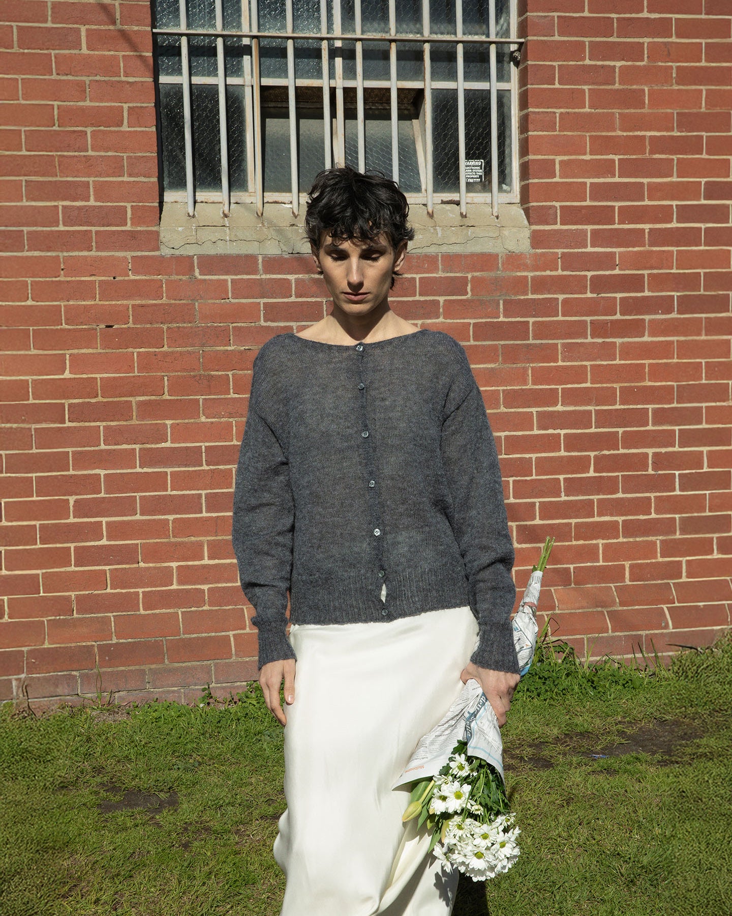 A person stands in front of a brick wall, wearing Francie's relaxed Fleur Cardigan in Shadow Grey and a white skirt, while holding a bouquet of white flowers.