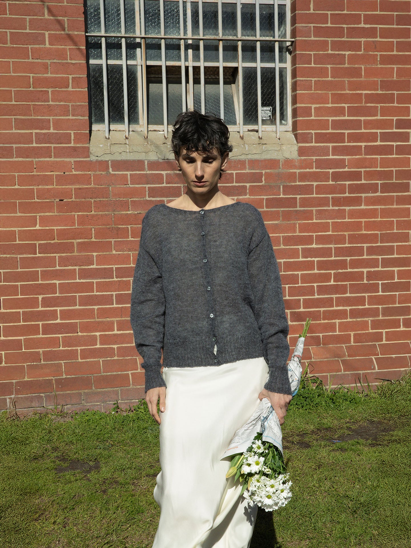 A person stands in front of a brick wall, wearing Francie's relaxed Fleur Cardigan in Shadow Grey and a white skirt, while holding a bouquet of white flowers.
