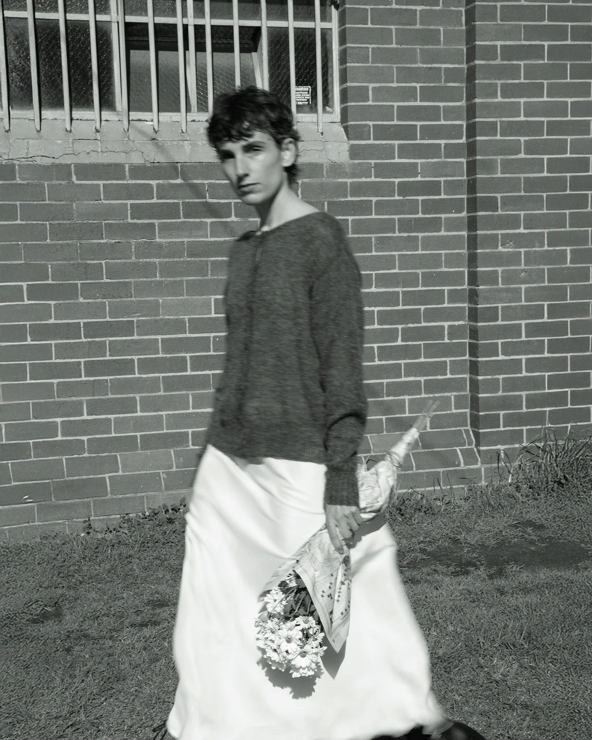 A calm individual wearing the Francie Fleur Cardigan in Shadow Grey and a long skirt strolls past a brick wall, holding a bouquet wrapped in paper.