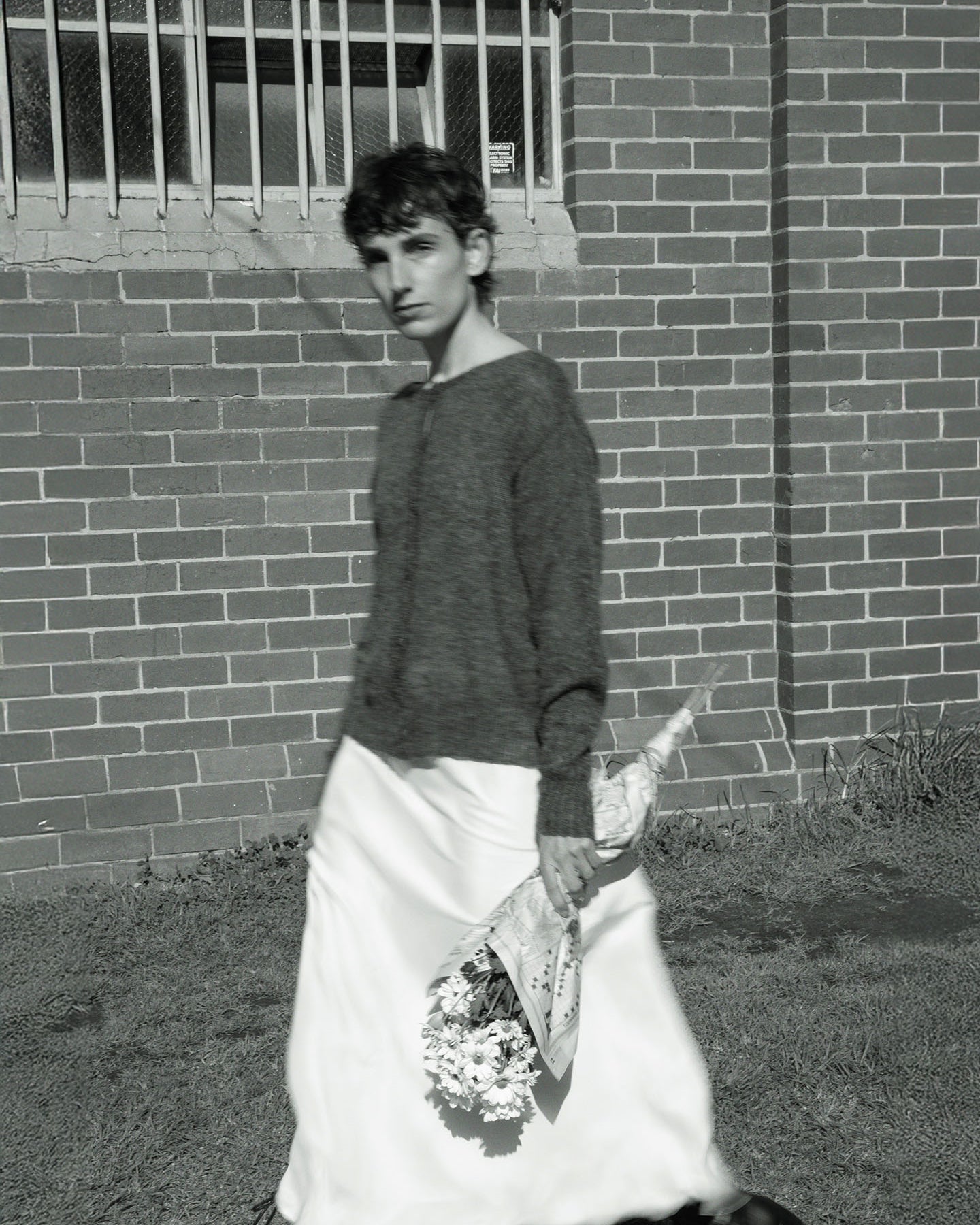 A person stands in front of a brick wall, wearing Francie's relaxed Fleur Cardigan in Shadow Grey and a white skirt, while holding a bouquet of white flowers.