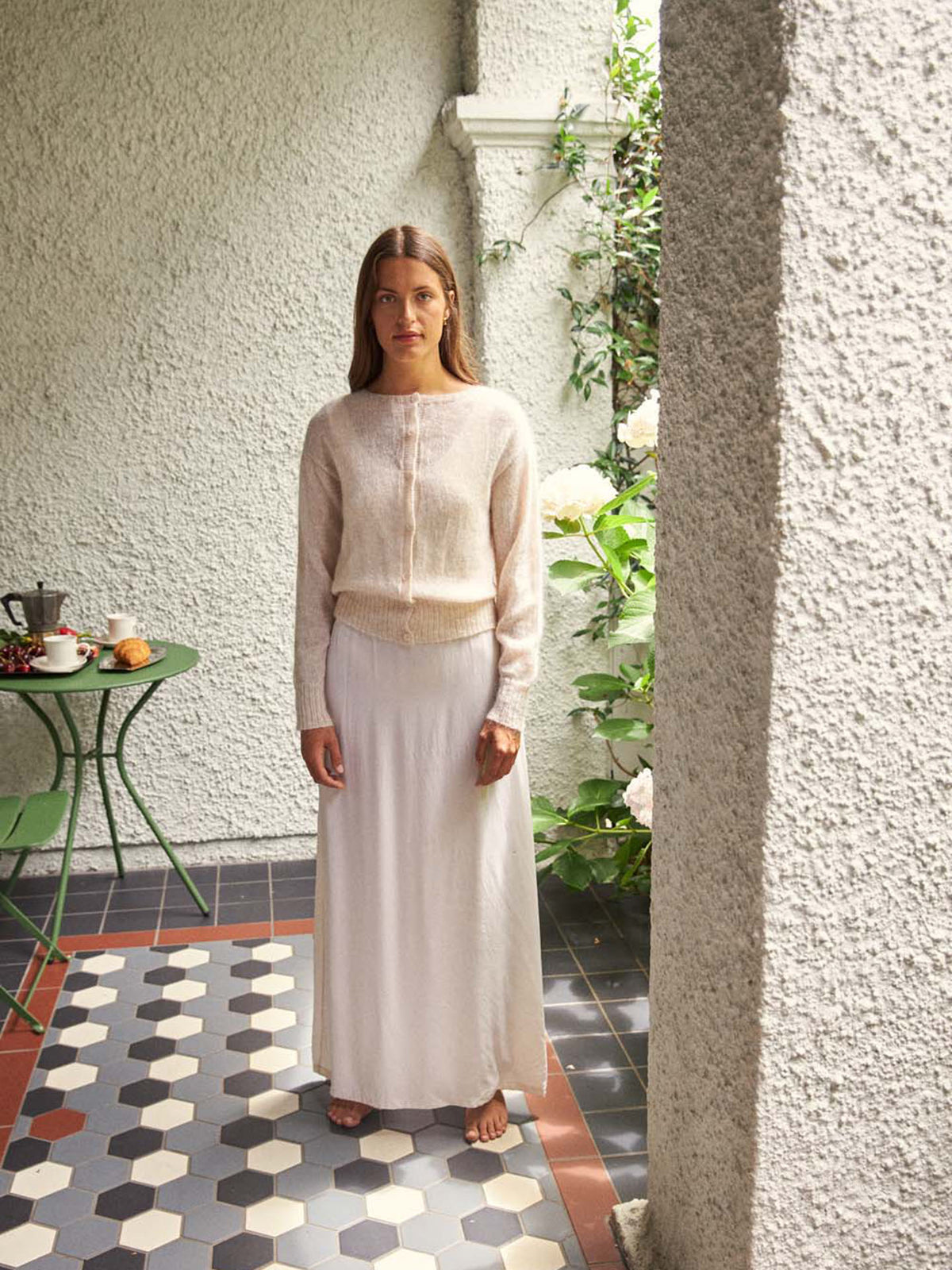 A petite woman in the Fleur Cardigan – Creme, a relaxed light-knit sweater, and a long white skirt stands barefoot on a tiled patio beside a green table set with food and drink.