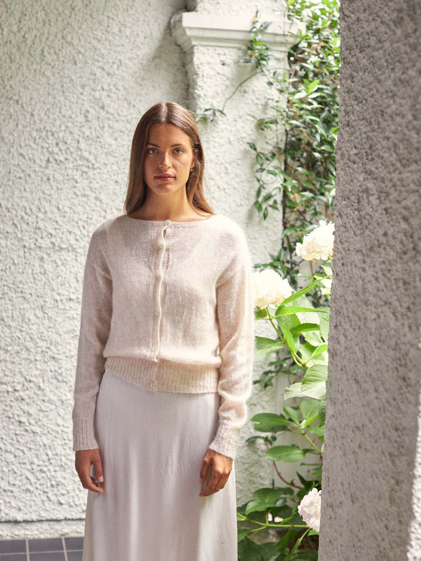 A woman wearing the relaxed fit Fleur Cardigan in Creme stands in front of a textured wall and green plants with white flowers.