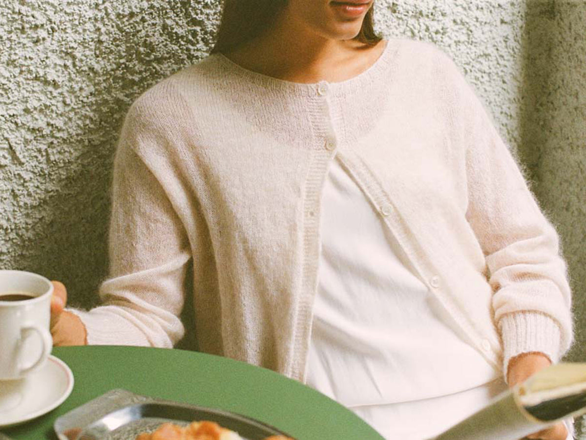 A petite person wearing the Fleur Cardigan – Creme sits at a green table with coffee and a pastry, reading a newspaper.