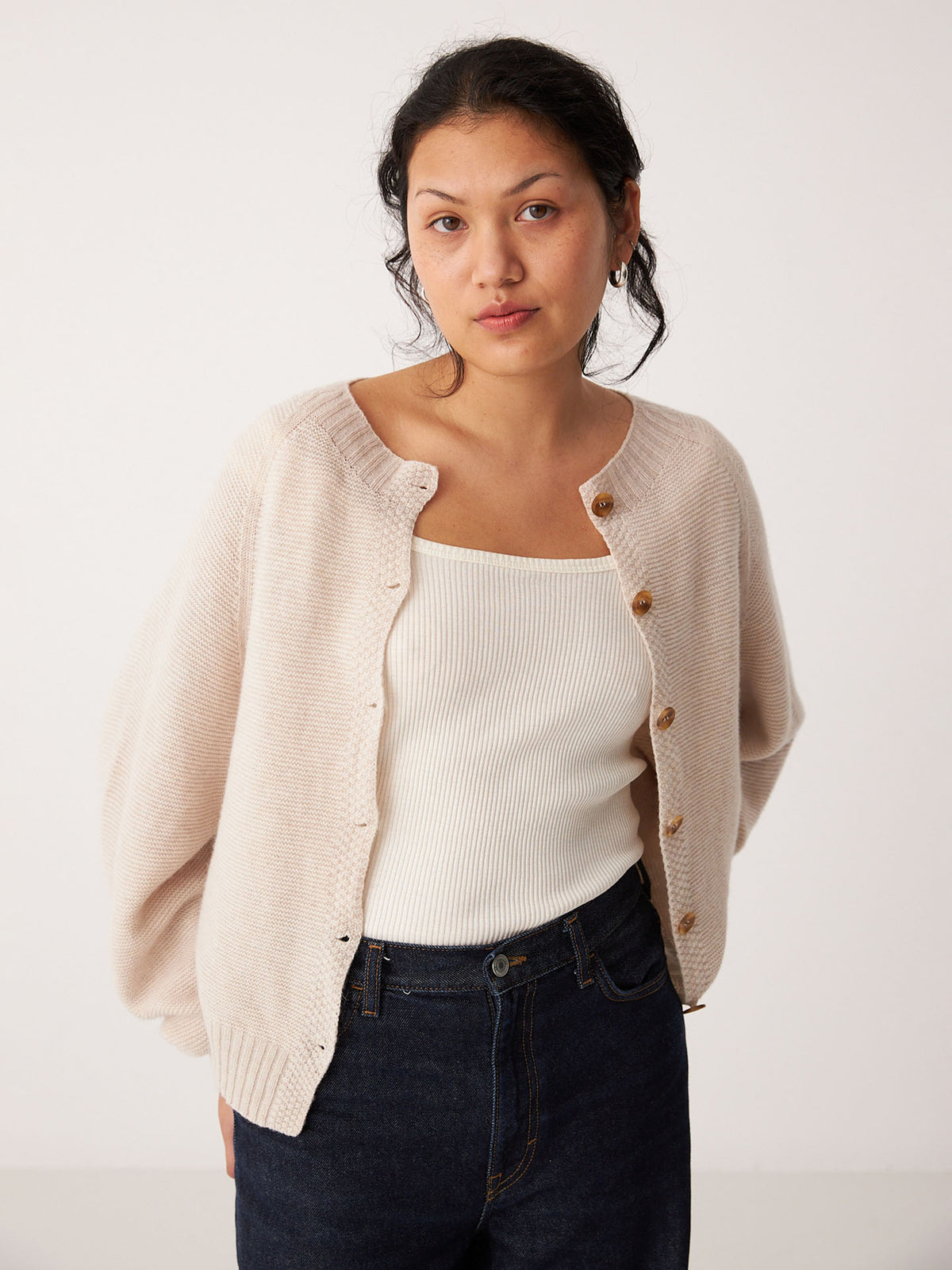 A woman in a Francie Garter Loft Cardigan – Creme, worn oversized with one side unbuttoned, pairs it with a white ribbed tank top and dark jeans while standing against a plain white background.