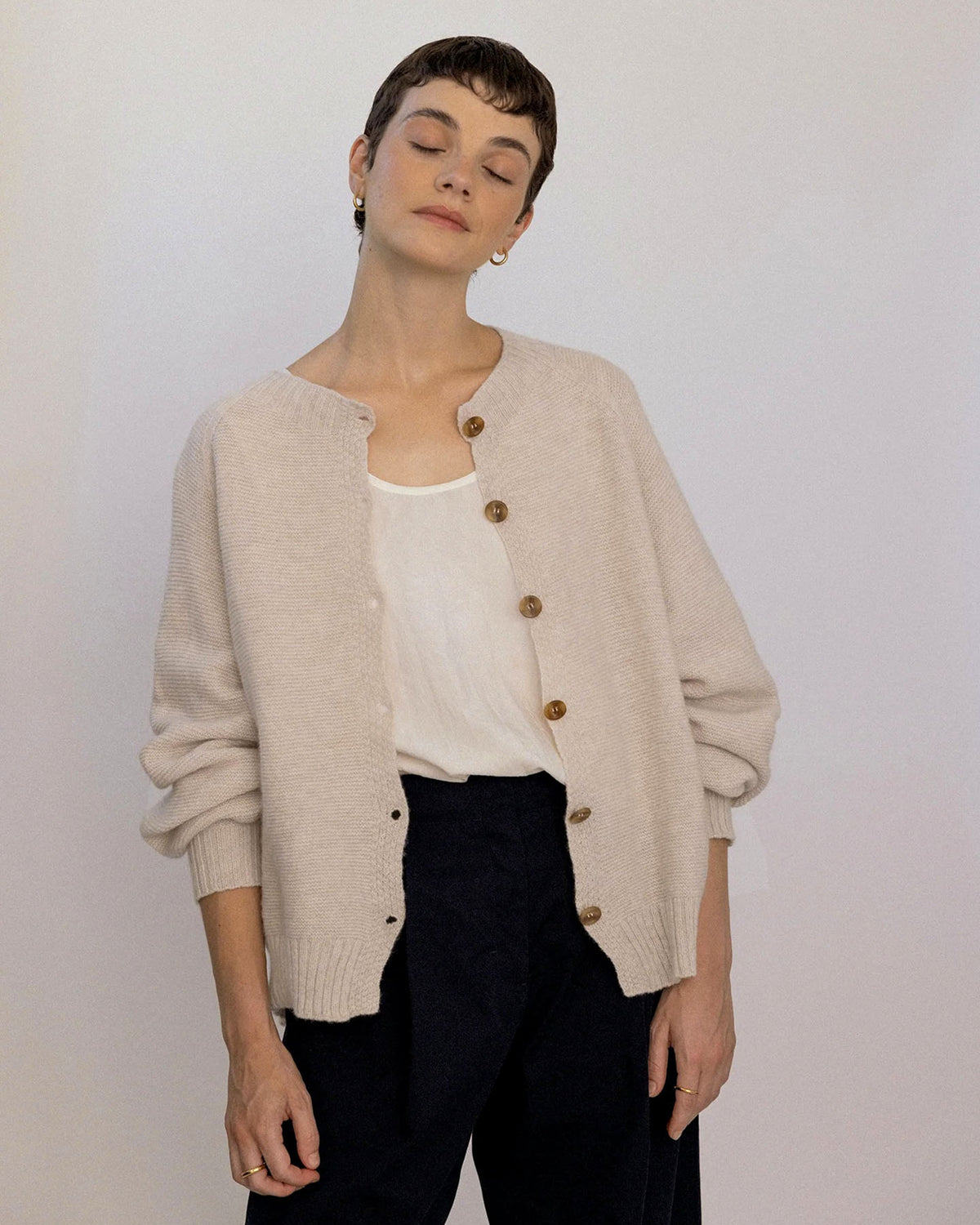 A person with short hair wears the Loft Cardigan – Creme over a white top and black pants, standing against a plain light background with eyes closed.