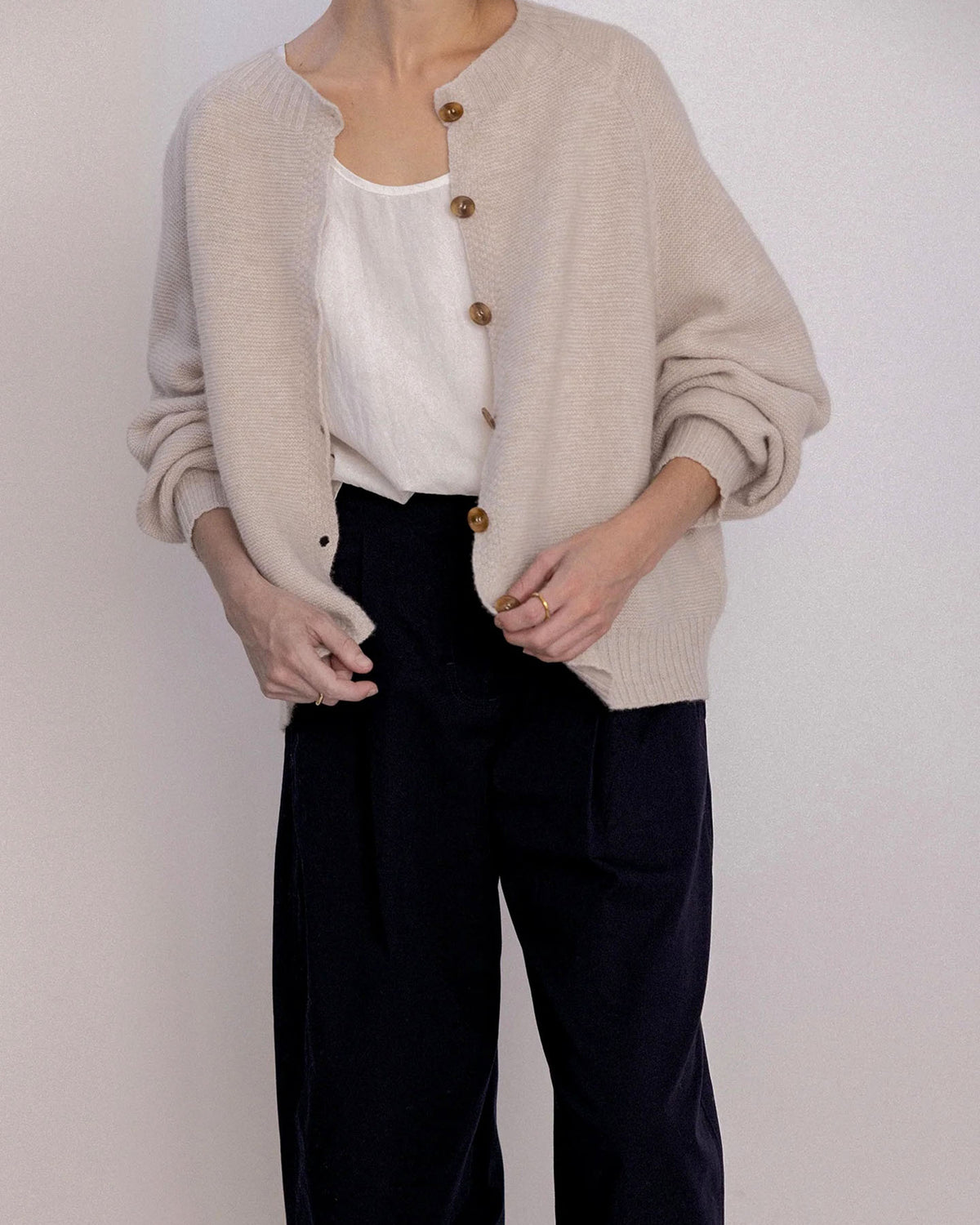 A person stands against a plain background with hands near the buttons, wearing a Loft Cardigan – Creme over a white top and black pants.