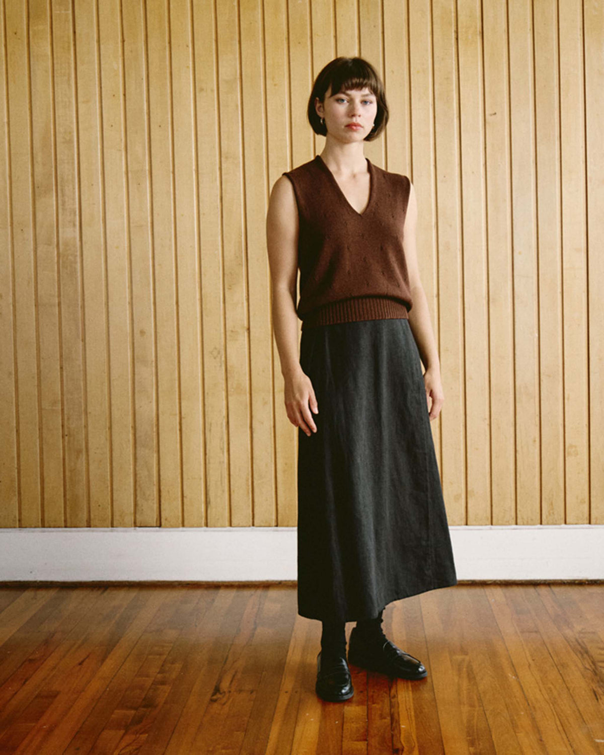 Person standing on a wooden floor against a wood-paneled wall, wearing a sleeveless brown sweater, long black skirt, and black shoes.