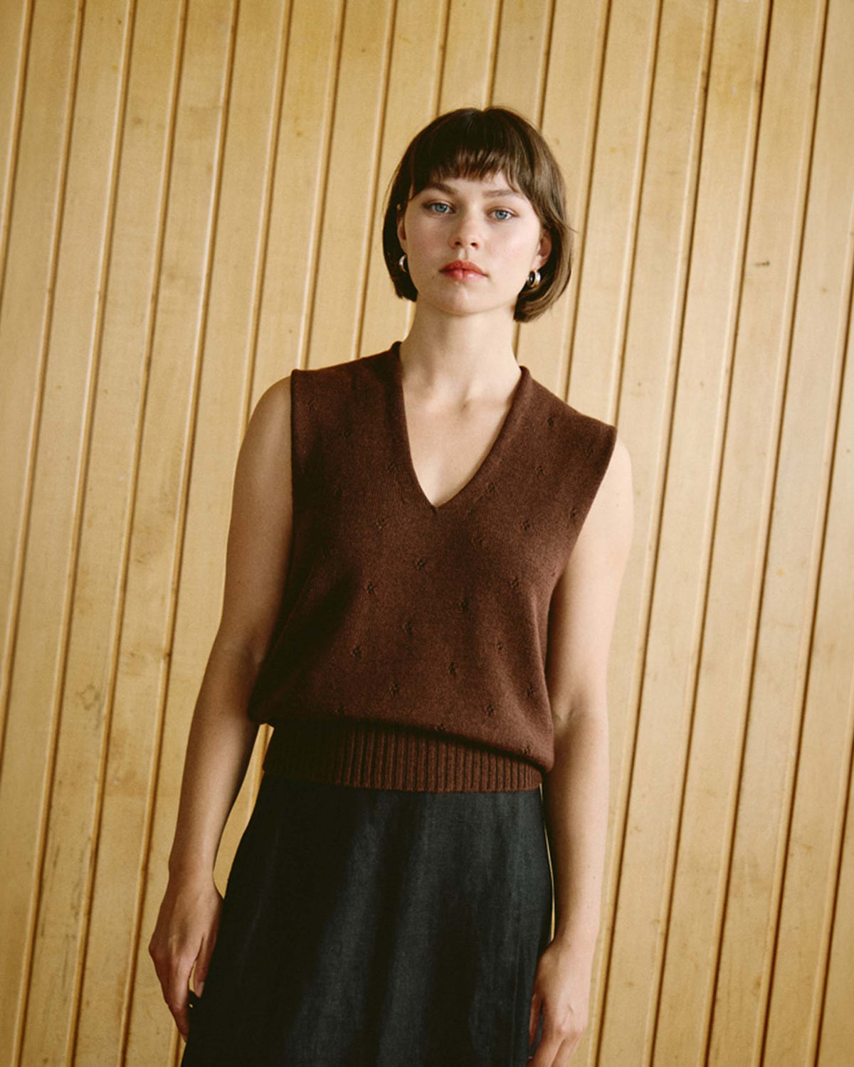 Woman with short dark hair wears a sleeveless brown V-neck sweater and dark skirt, standing in front of a wooden panel wall.