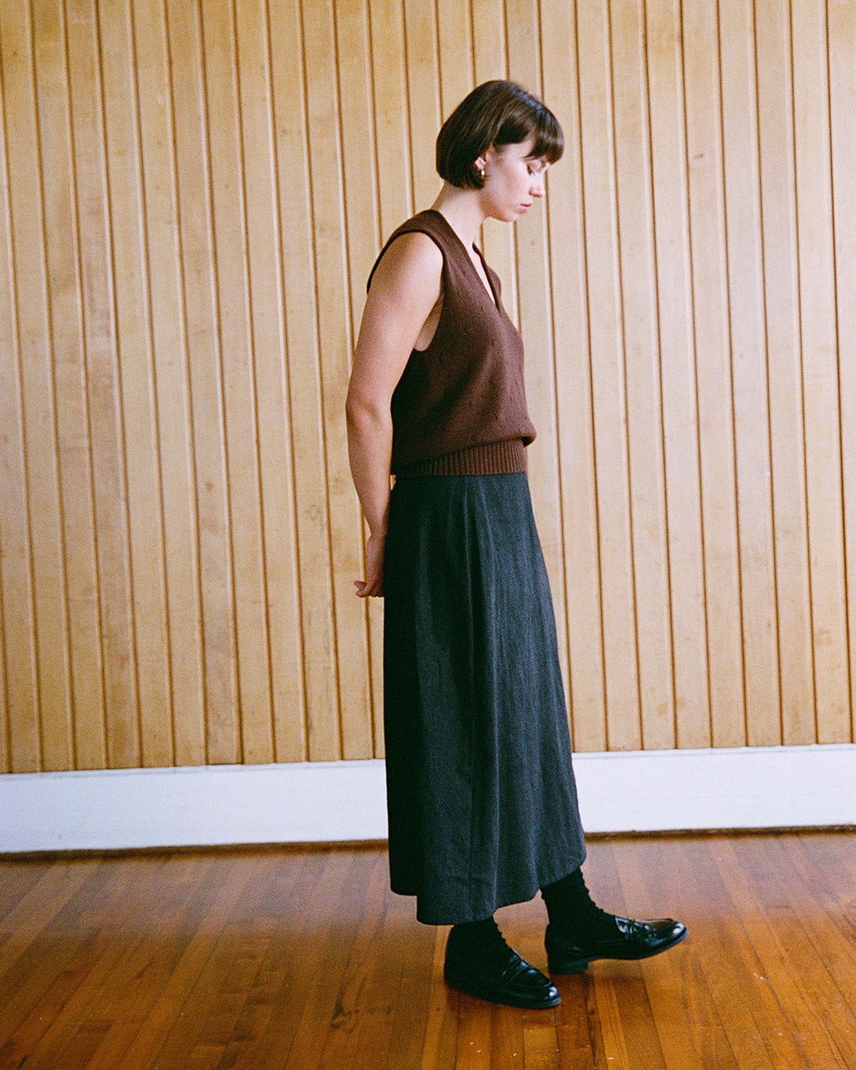 A person with short hair stands sideways in a room with wooden floors and a vertical wood-paneled wall, wearing a brown sleeveless top, dark skirt, and black shoes.