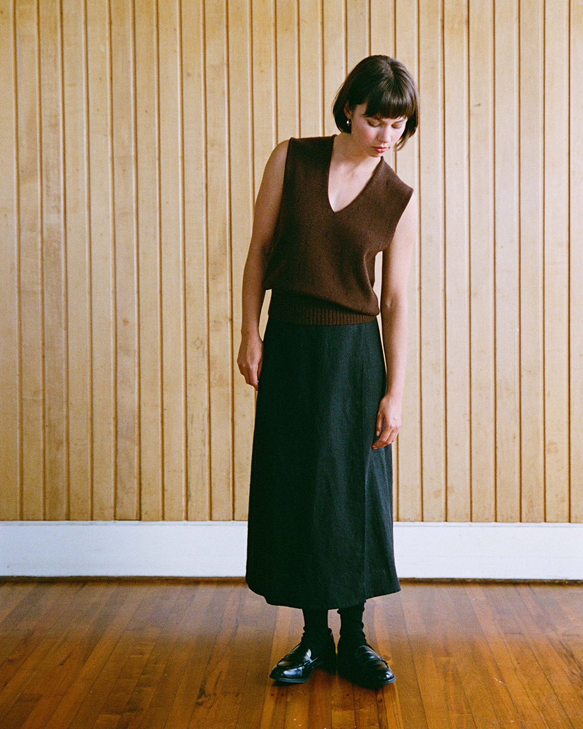 A woman wearing a sleeveless brown sweater and long black skirt stands indoors against a wooden panel wall, looking down to her left.