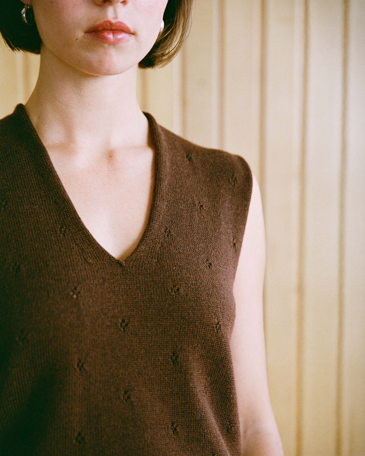 A person with short hair wearing a brown sleeveless knit top stands in front of a light wooden wall. Their face is partially visible, showing a neutral expression.