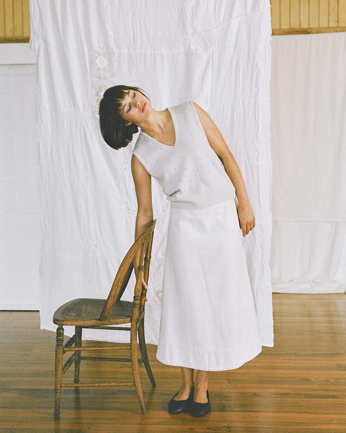 A woman in a white sleeveless top and skirt leans her head on the back of a wooden chair, standing on a wooden floor with white fabric hanging in the background.