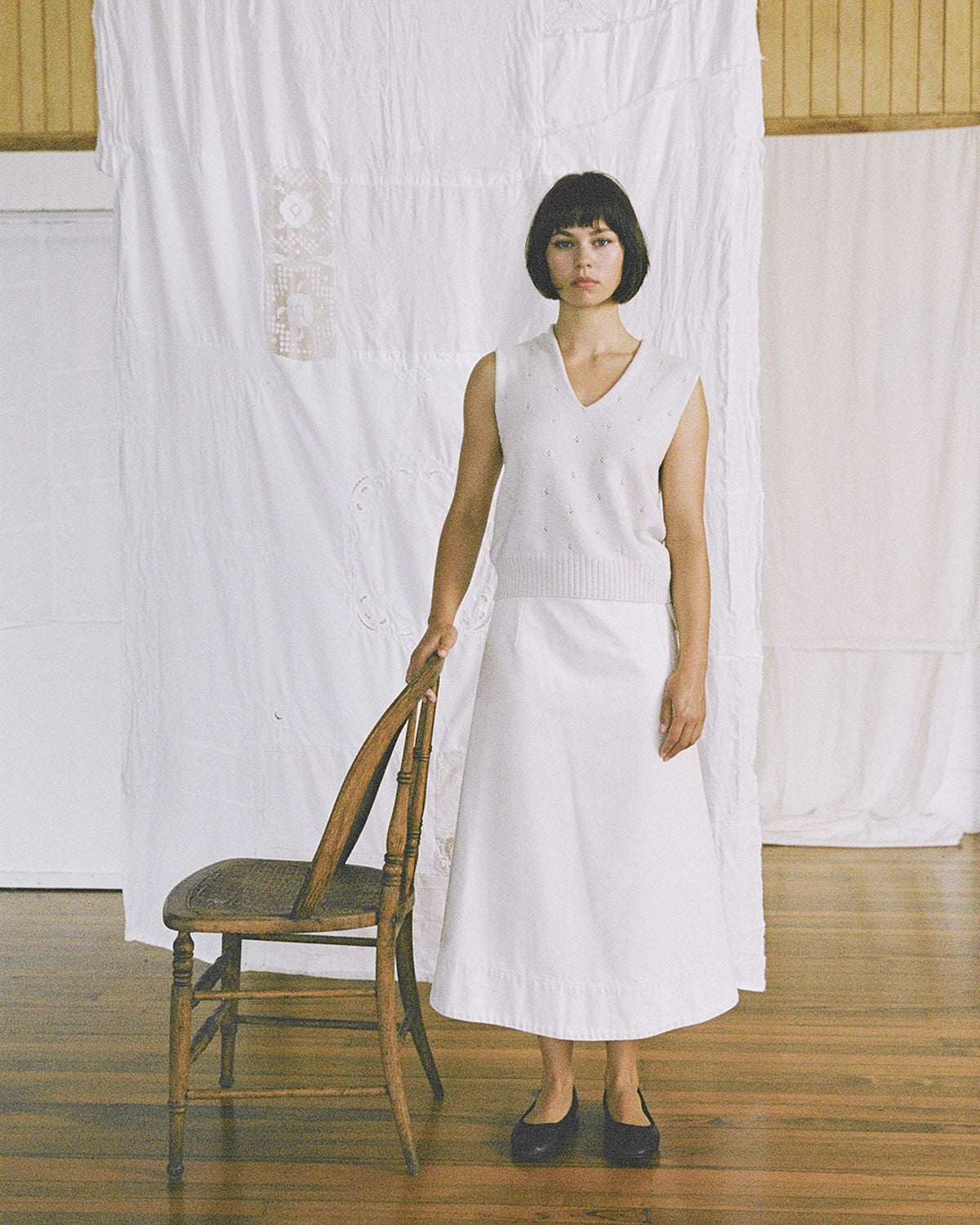 A woman in a sleeveless white top and long white skirt stands next to a wooden chair on a wooden floor, with white fabric draped in the background.