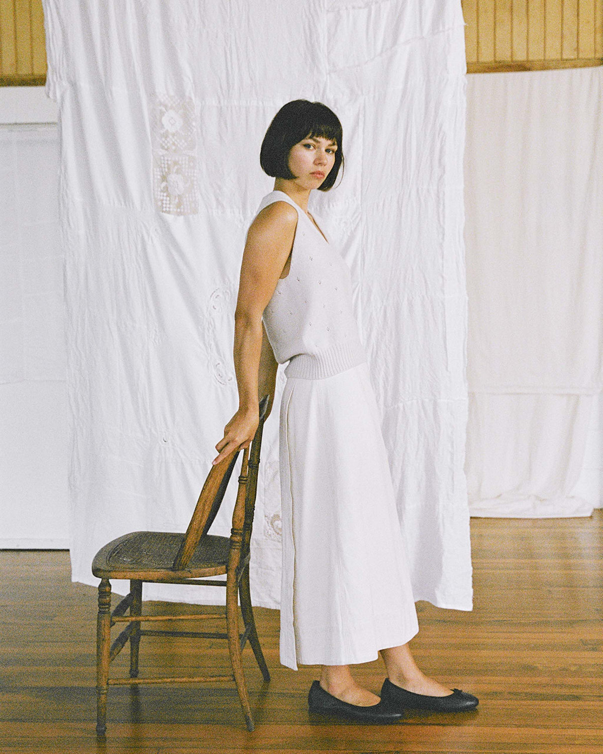 Woman in a white sleeveless top and skirt stands sideways next to a wooden chair, with a white sheet hanging in the background on a wooden floor.