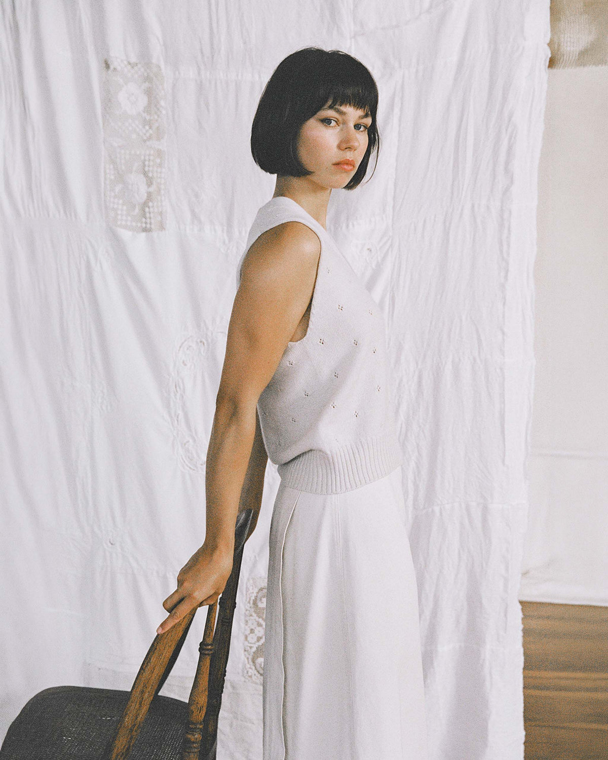 A person with short dark hair stands sideways, holding the back of a wooden chair, wearing a sleeveless white top and long white skirt, in front of a white fabric backdrop.