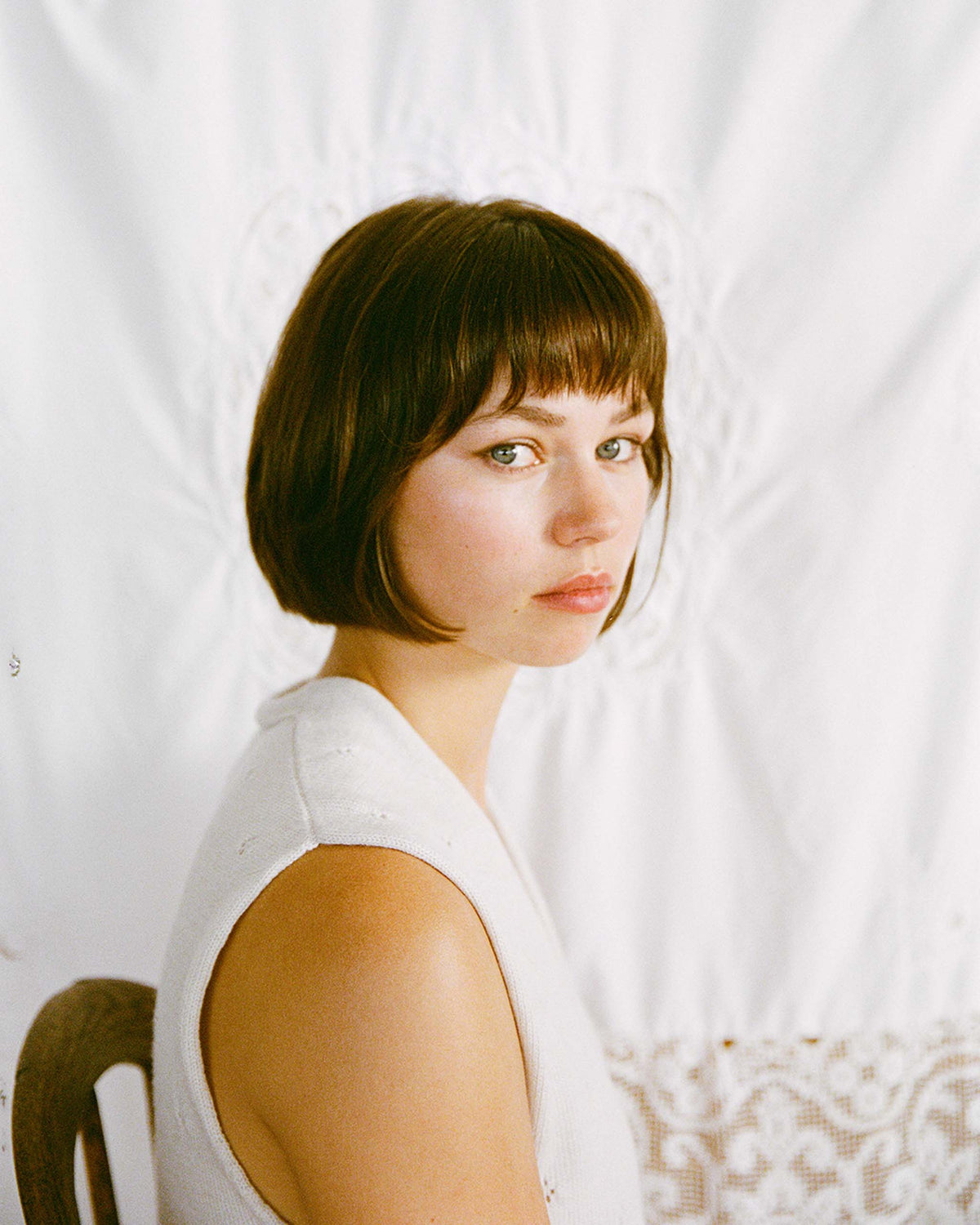 A young woman with short brown hair and bangs sits sideways on a chair, looking at the camera, with a white patterned fabric backdrop behind her.