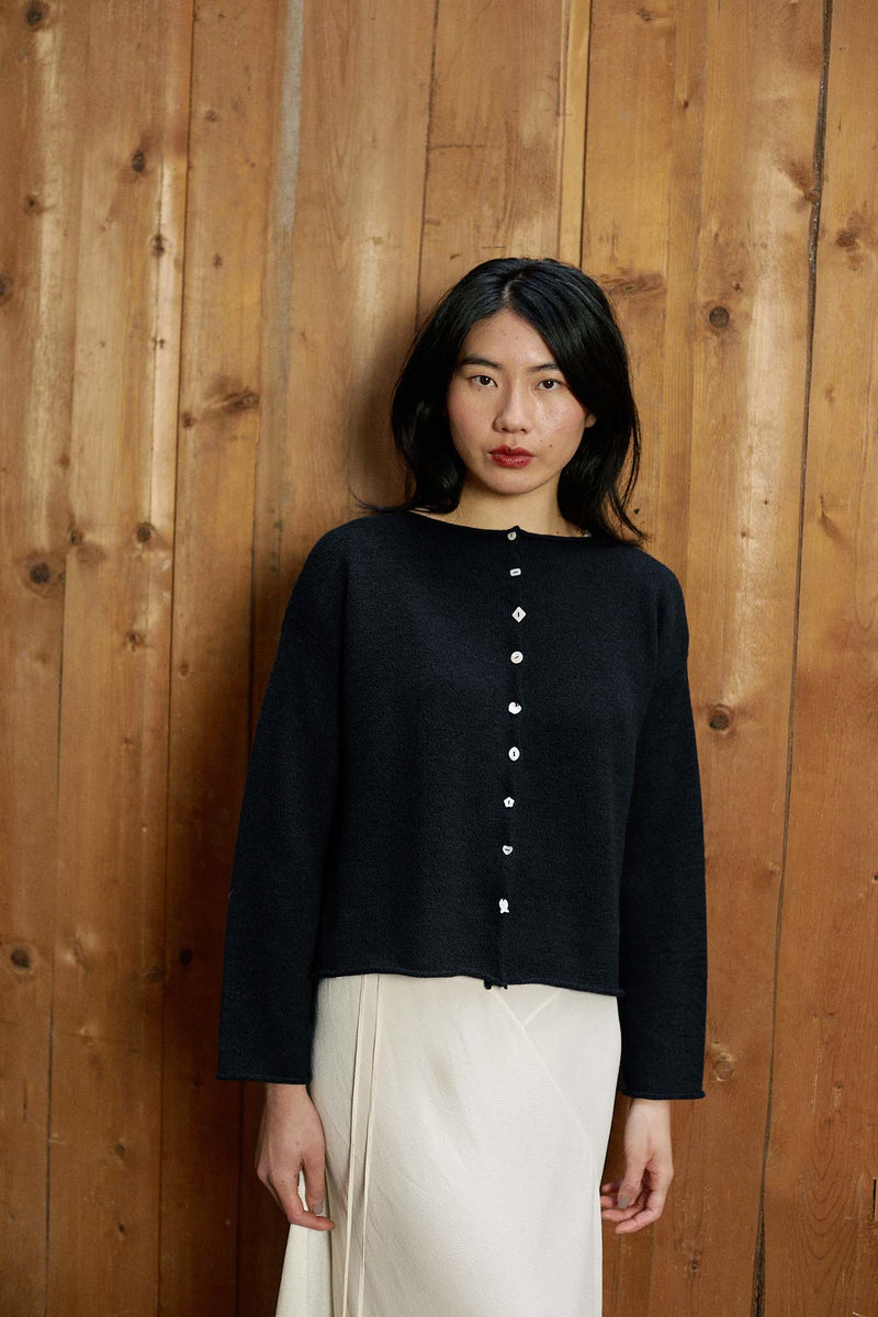 A woman stands before a wooden wall, wearing the Heirloom Doll's Cardigan in black with a relaxed fit and a cream skirt, gazing at the camera with a neutral expression.
