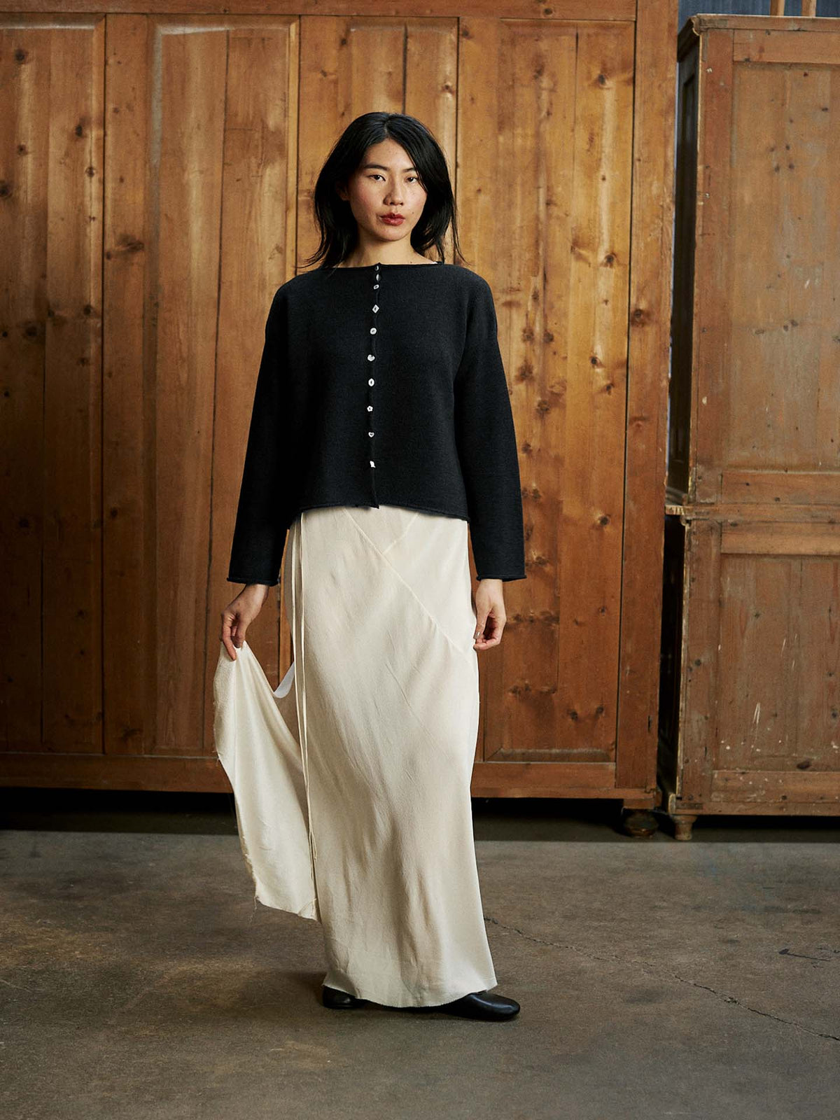 A woman stands indoors against wooden panels, wearing the Heirloom Doll's Cardigan in black with a relaxed fit and long sleeves, paired with a flowing white skirt and holding the skirt slightly with one hand.