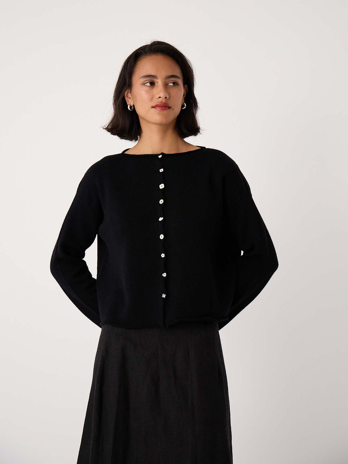 A woman wears the Heirloom Doll's Cardigan – Black, a relaxed fit black cardigan with white buttons, paired with a dark skirt against a plain light background.