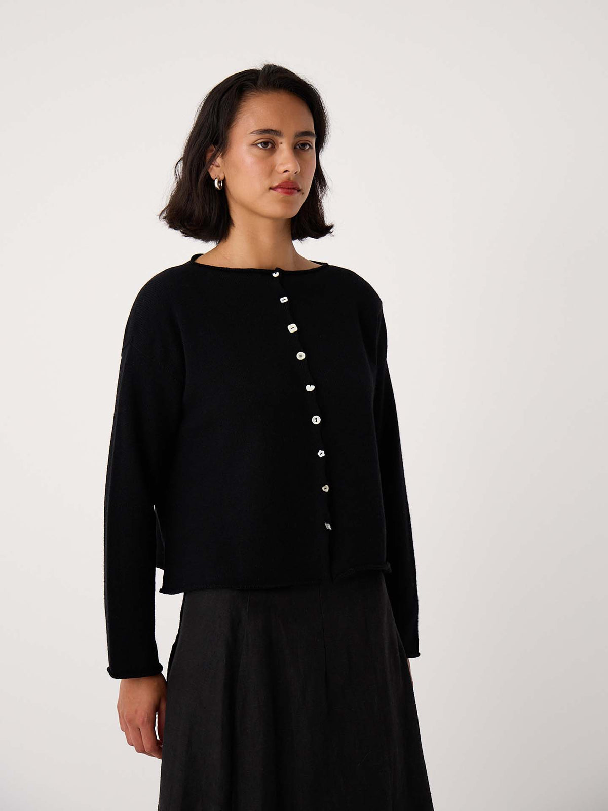 A person wears the Heirloom Doll's Cardigan in black, featuring a relaxed fit and white buttons, paired with a black skirt against a plain light background.