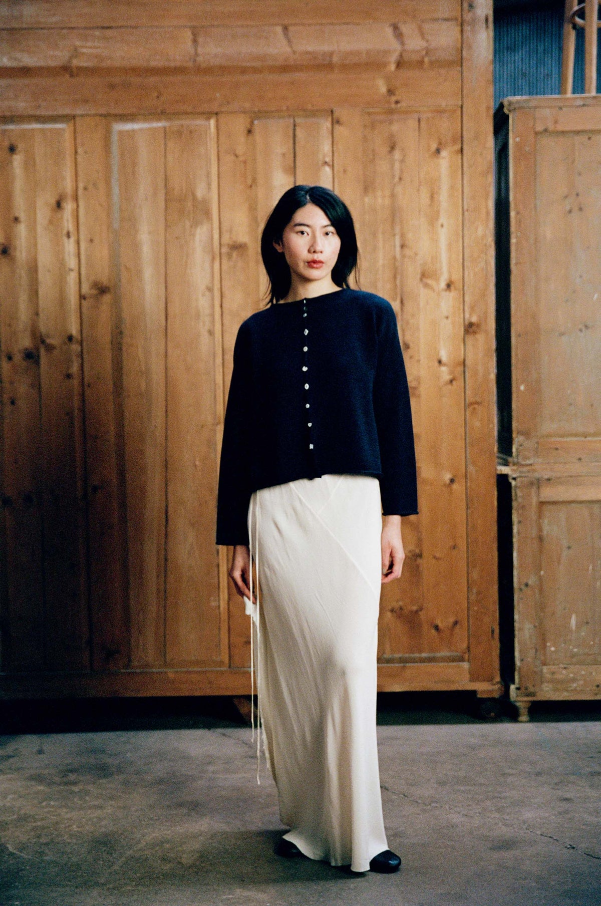 A woman stands indoors wearing the Heirloom Doll's Cardigan in Ink, styled in a relaxed fit with a long white skirt, against a backdrop of wooden panels and cabinets.