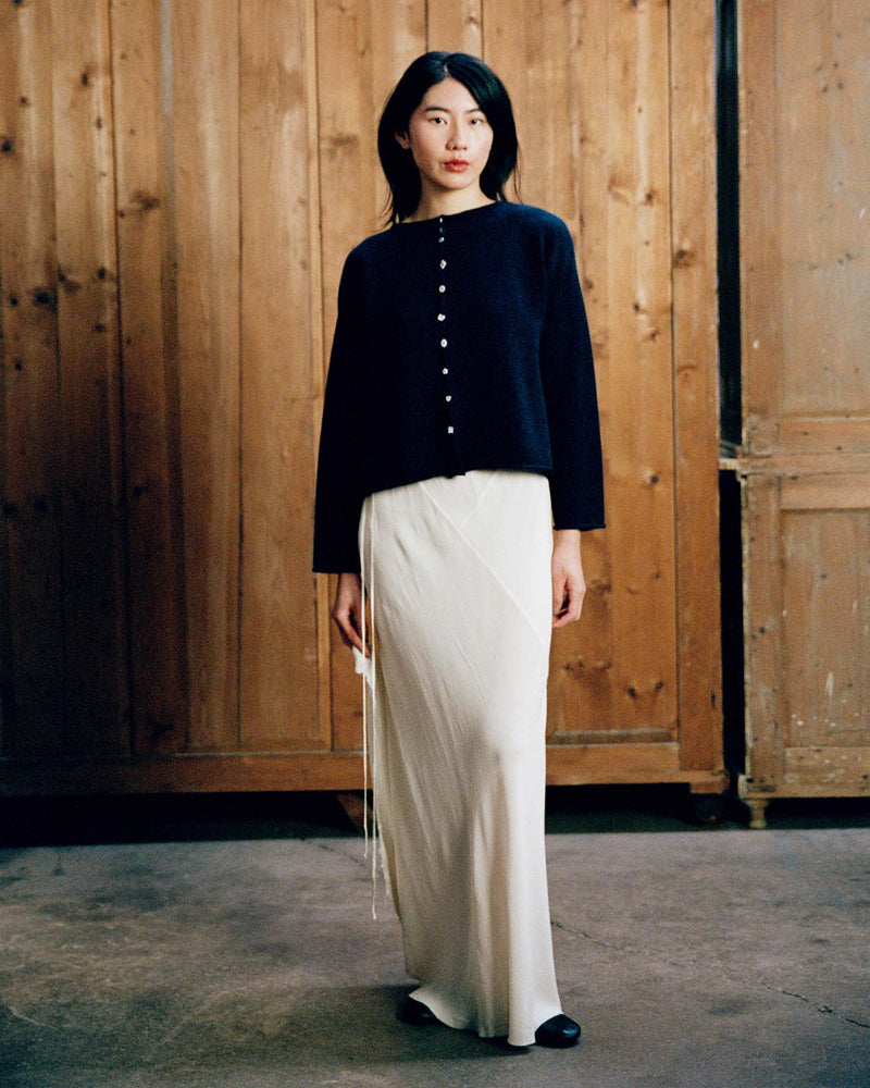 A woman stands indoors wearing the Heirloom Doll's Cardigan in Ink, styled in a relaxed fit with a long white skirt, against a backdrop of wooden panels and cabinets.