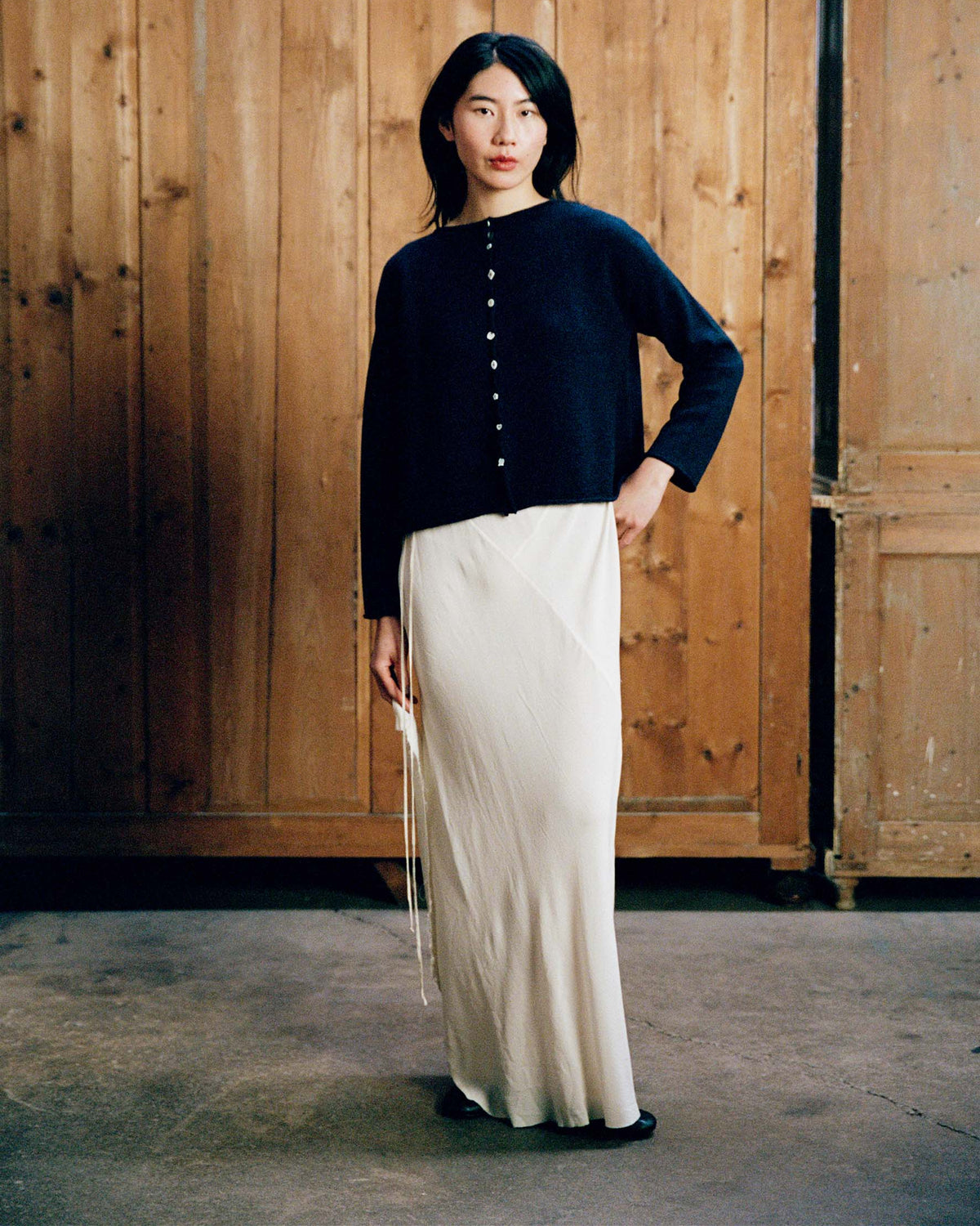 A woman stands indoors before wooden panels, wearing the Heirloom Doll's Cardigan – Ink in a petite cropped style with buttons, paired with a long white skirt.