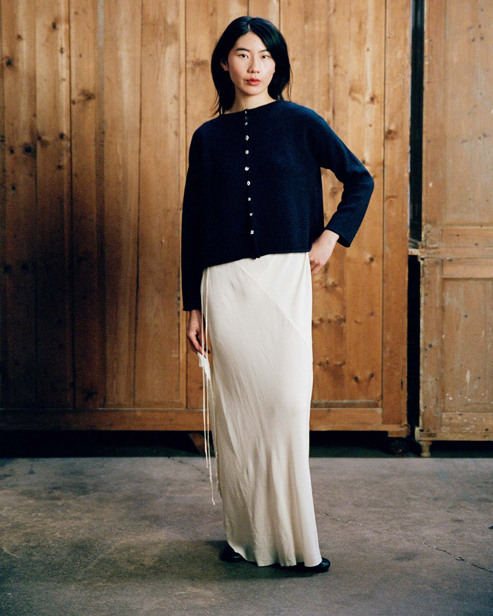 A woman stands indoors wearing the Heirloom Doll's Cardigan in Ink, styled in a relaxed fit with a long white skirt, against a backdrop of wooden panels and cabinets.