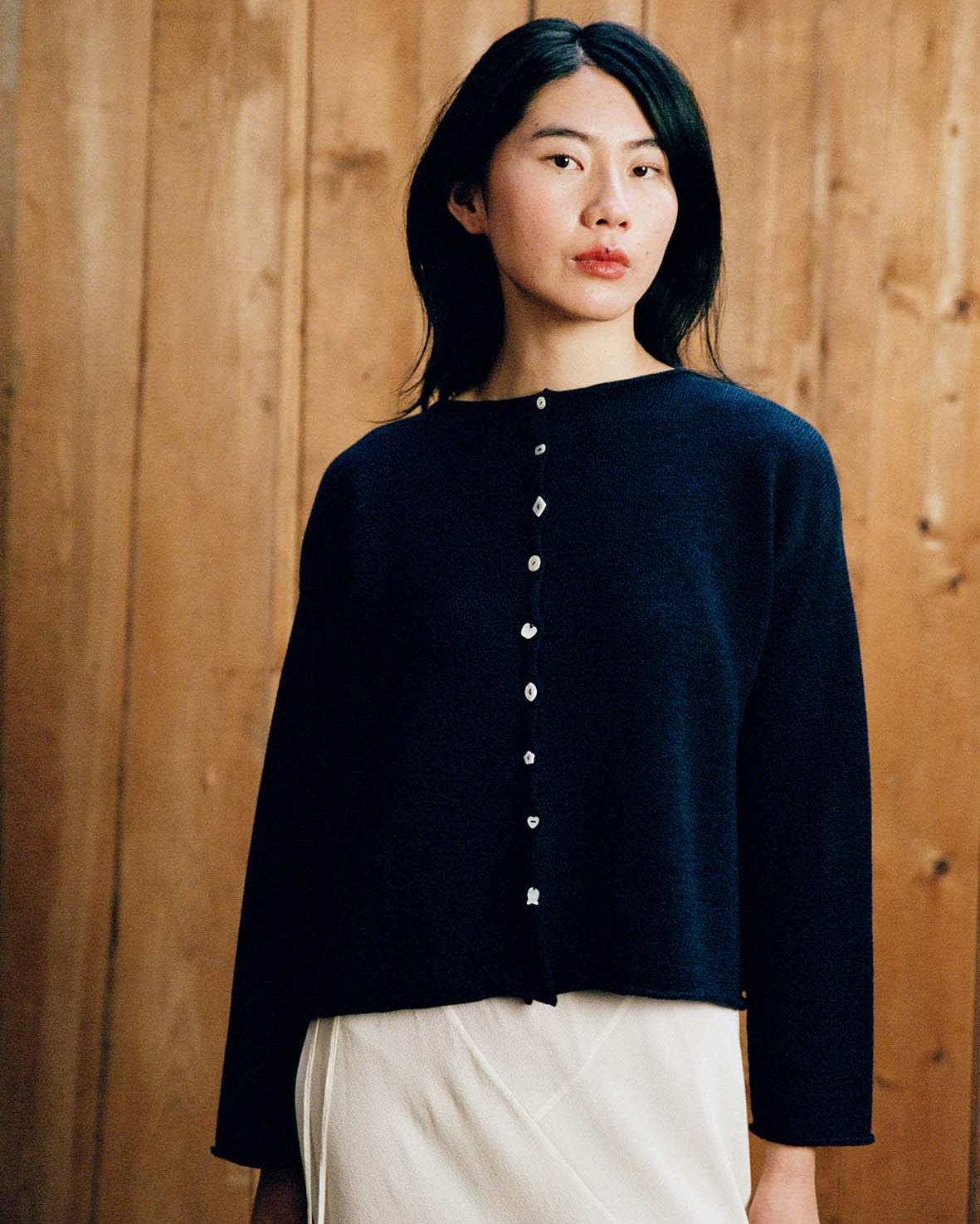 A person with long dark hair is wearing the Heirloom Doll's Cardigan – Ink, a relaxed-fit navy sweater, over a light skirt while standing in front of a wooden wall.