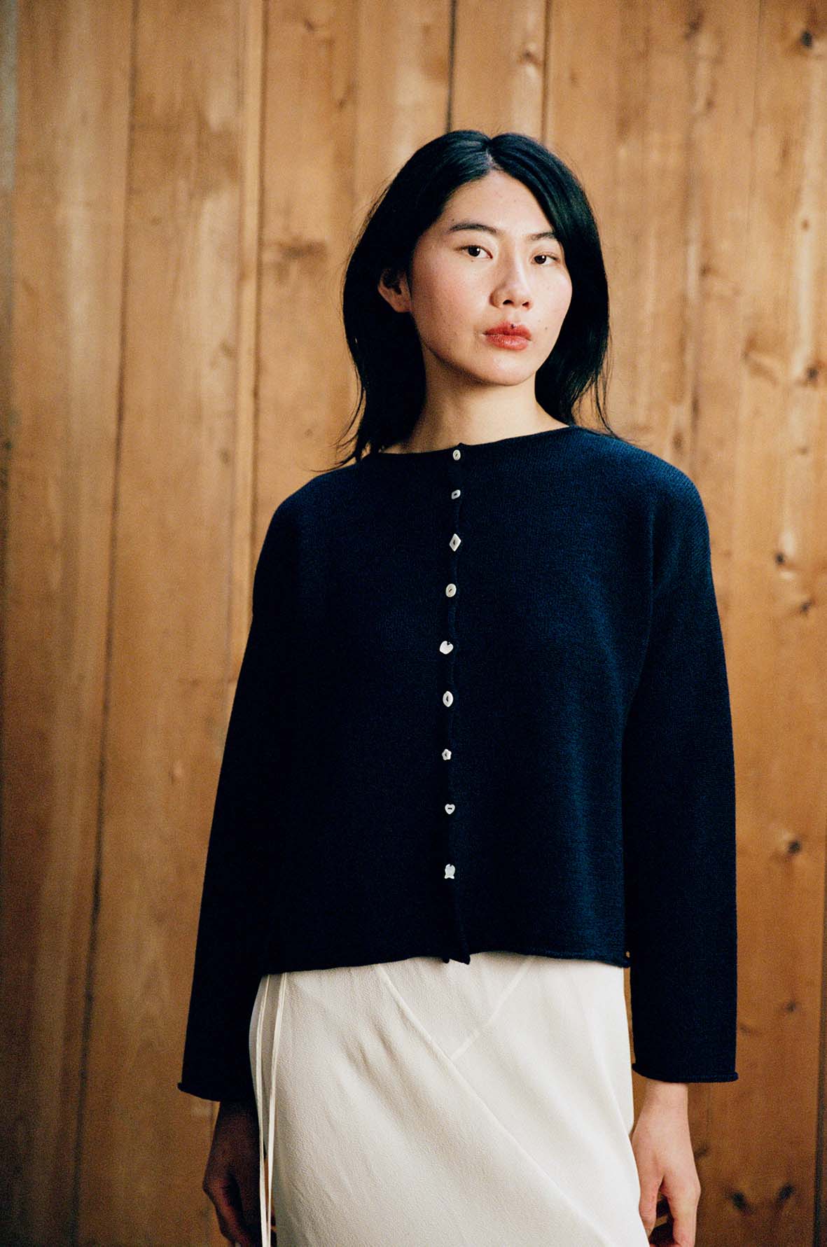 A person with long dark hair is wearing the Heirloom Doll's Cardigan – Ink, a relaxed-fit navy sweater, over a light skirt while standing in front of a wooden wall.
