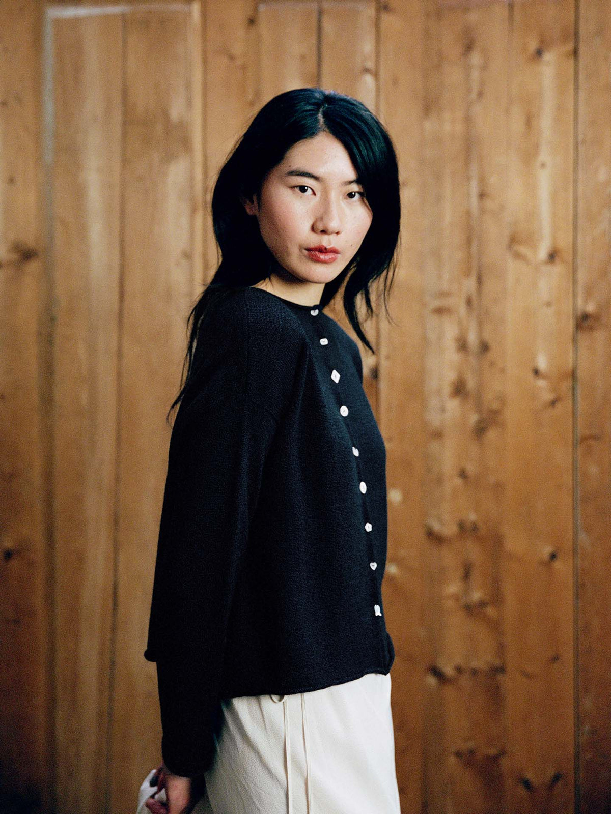 A woman with long black hair models the Heirloom Doll's Cardigan – Ink in a petite cropped style with light-colored pants, standing in front of a wooden wall. Check our size guide for your perfect relaxed fit.