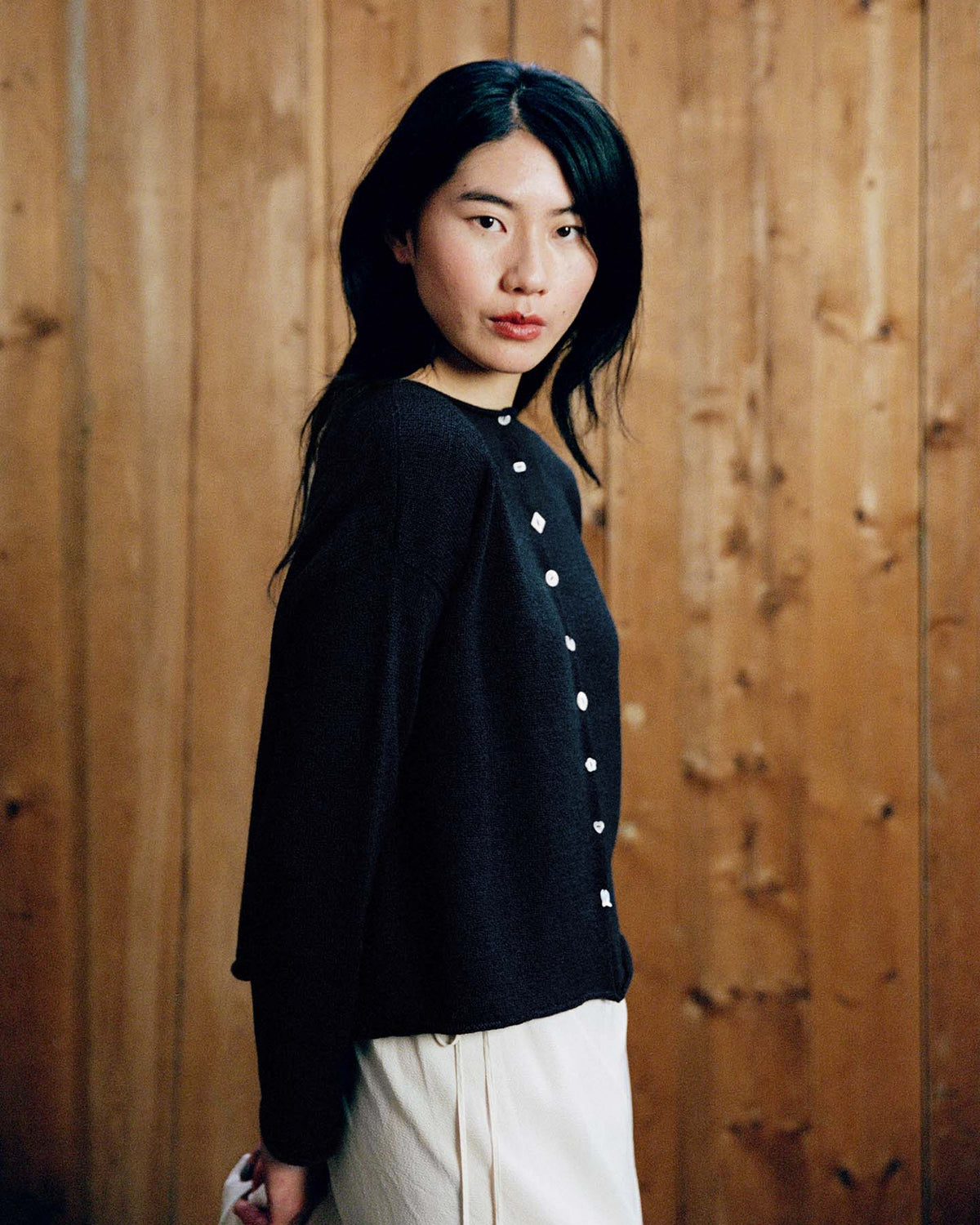 A woman with long black hair models the Heirloom Doll's Cardigan – Ink in a petite cropped style with light-colored pants, standing in front of a wooden wall. Check our size guide for your perfect relaxed fit.
