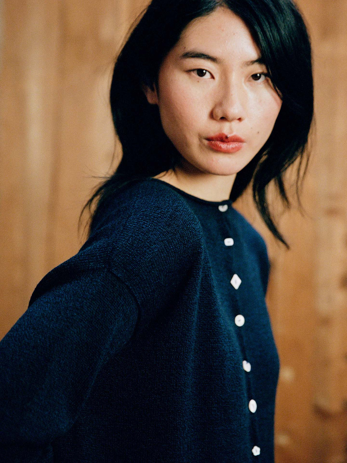 A woman with long black hair wears the Heirloom Doll's Cardigan – Ink, a navy blue buttoned sweater in a relaxed fit, standing before a wooden backdrop and gazing at the camera. Check our size guide for your perfect fit.