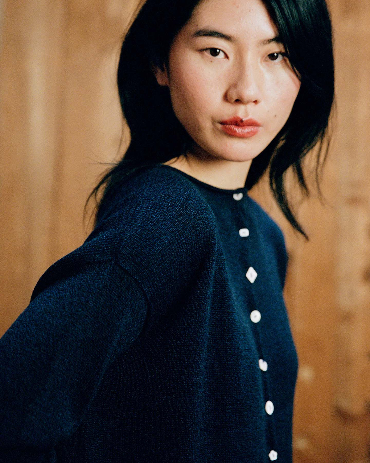 A woman with long black hair wears the Heirloom Doll's Cardigan – Ink, a navy blue buttoned sweater in a relaxed fit, standing before a wooden backdrop and gazing at the camera. Check our size guide for your perfect fit.