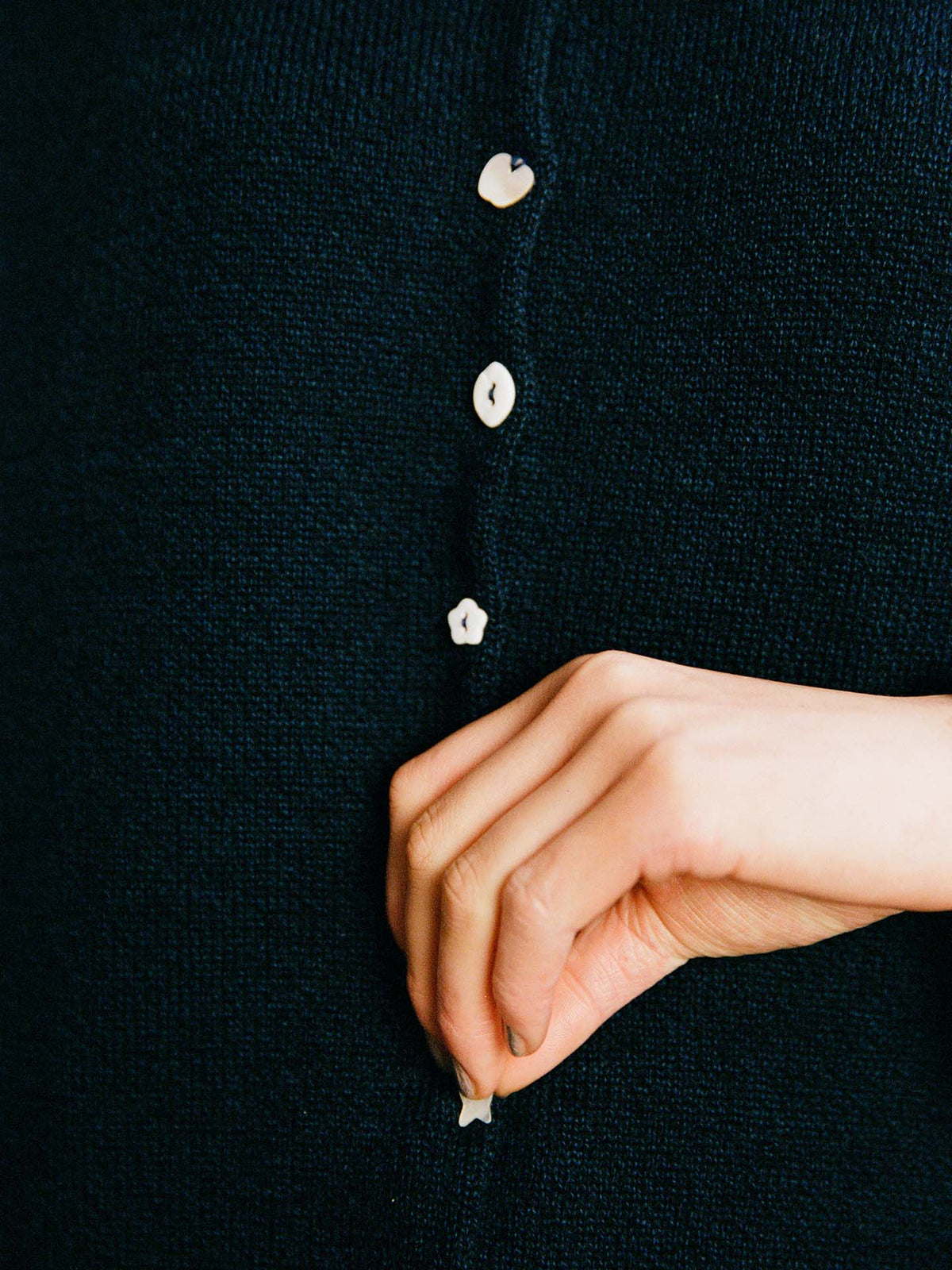 A hand buttons the Heirloom Doll's Cardigan in black, a dark knitted garment with small white buttons and a relaxed fit.