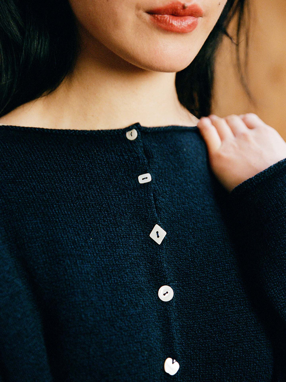 A woman wears the Heirloom Doll's Cardigan – Ink, a dark textured top with asymmetrical buttons in round, square, and heart shapes, showing off its relaxed fit as she holds the fabric near her shoulder.
