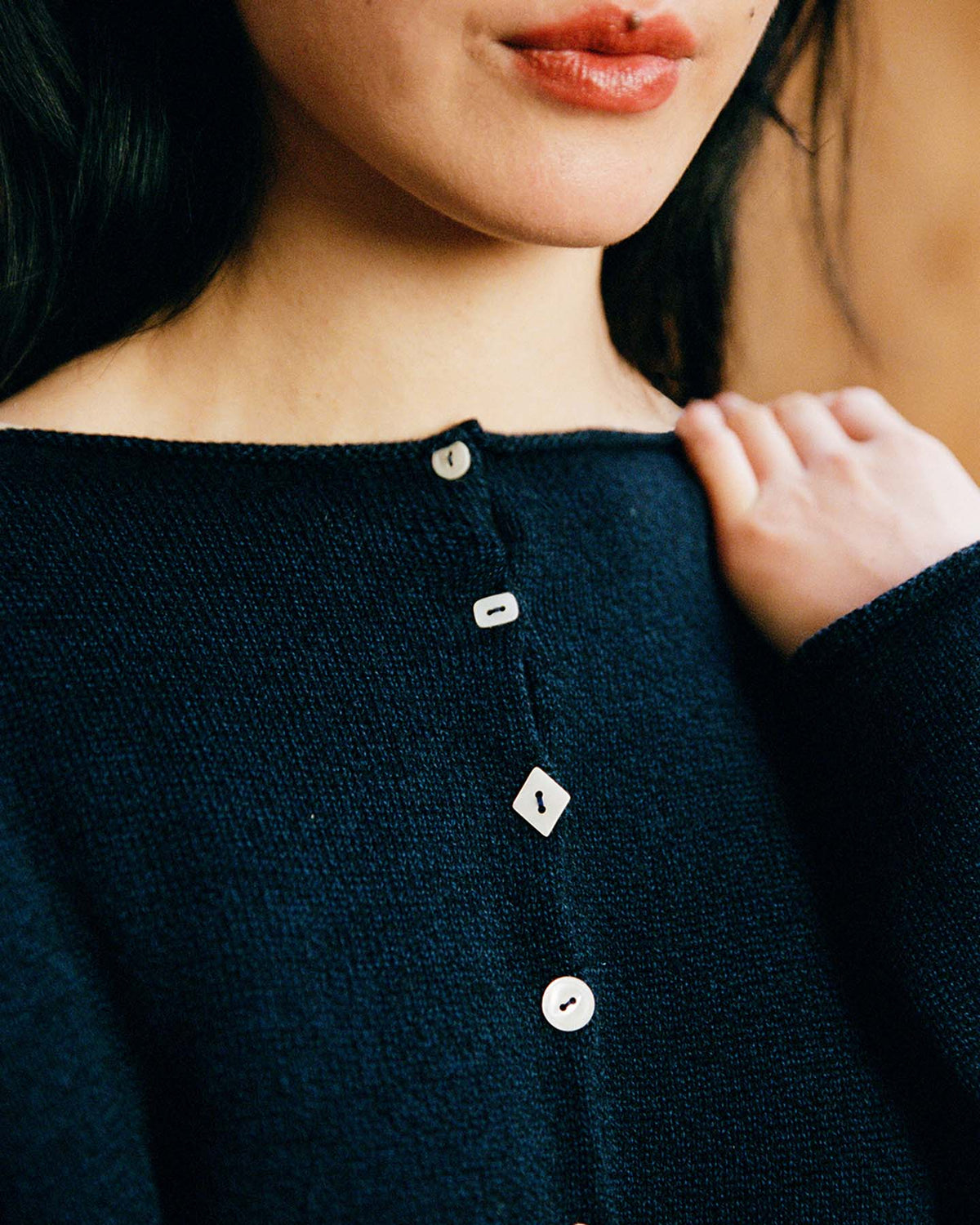 A woman wears the Heirloom Doll's Cardigan – Ink, a dark textured top with asymmetrical buttons in round, square, and heart shapes, showing off its relaxed fit as she holds the fabric near her shoulder.