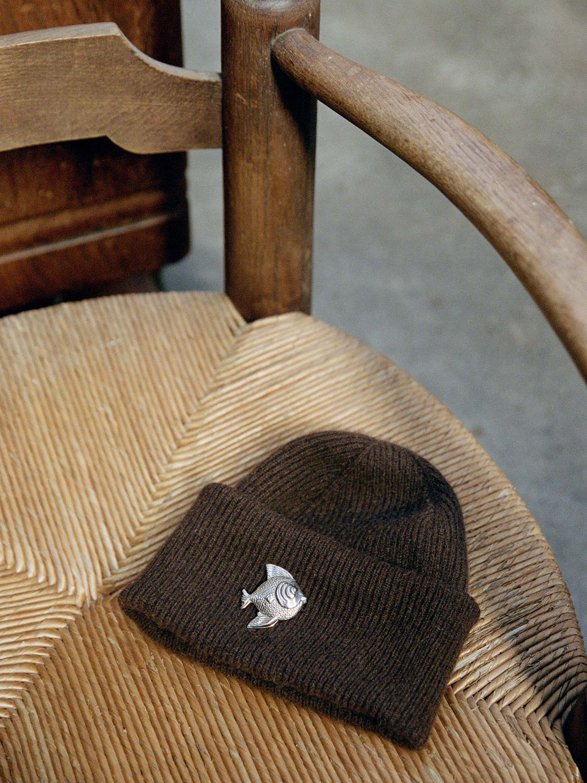 The Hinoki Beanie – Chestnut, featuring a metal fish pin, rests on a woven seat wooden chair.