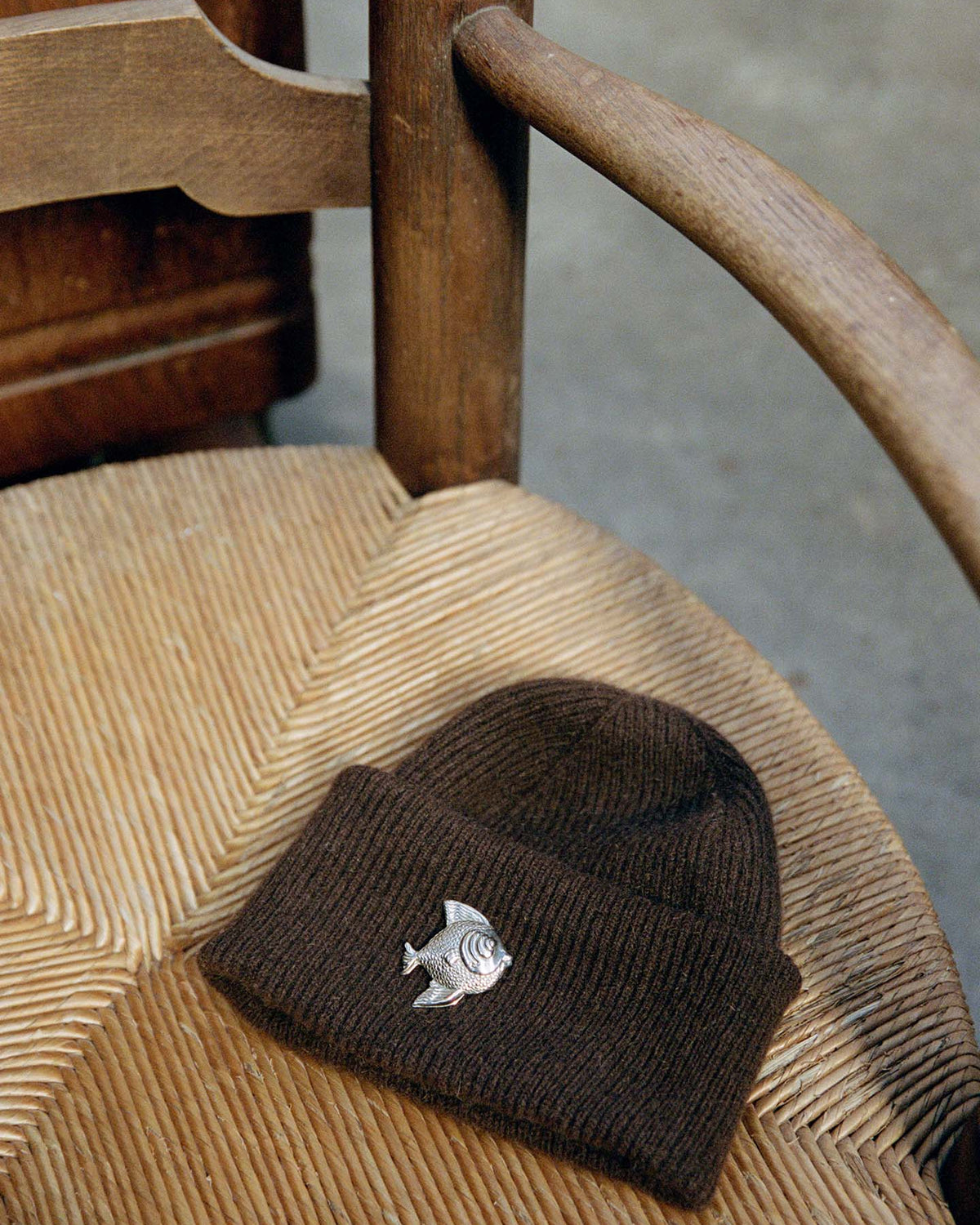 The Hinoki Beanie – Chestnut, featuring a metal fish pin, rests on a woven seat wooden chair.
