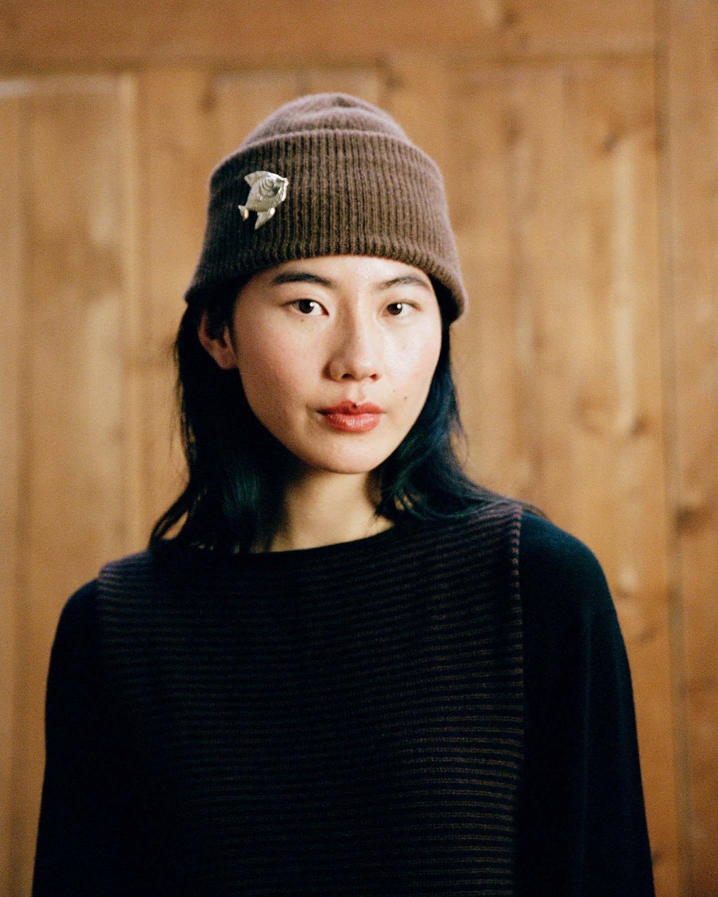 A person with shoulder-length dark hair wears the Hinoki Beanie – Chestnut, featuring a pin, and a dark striped top while standing in front of a wooden background.