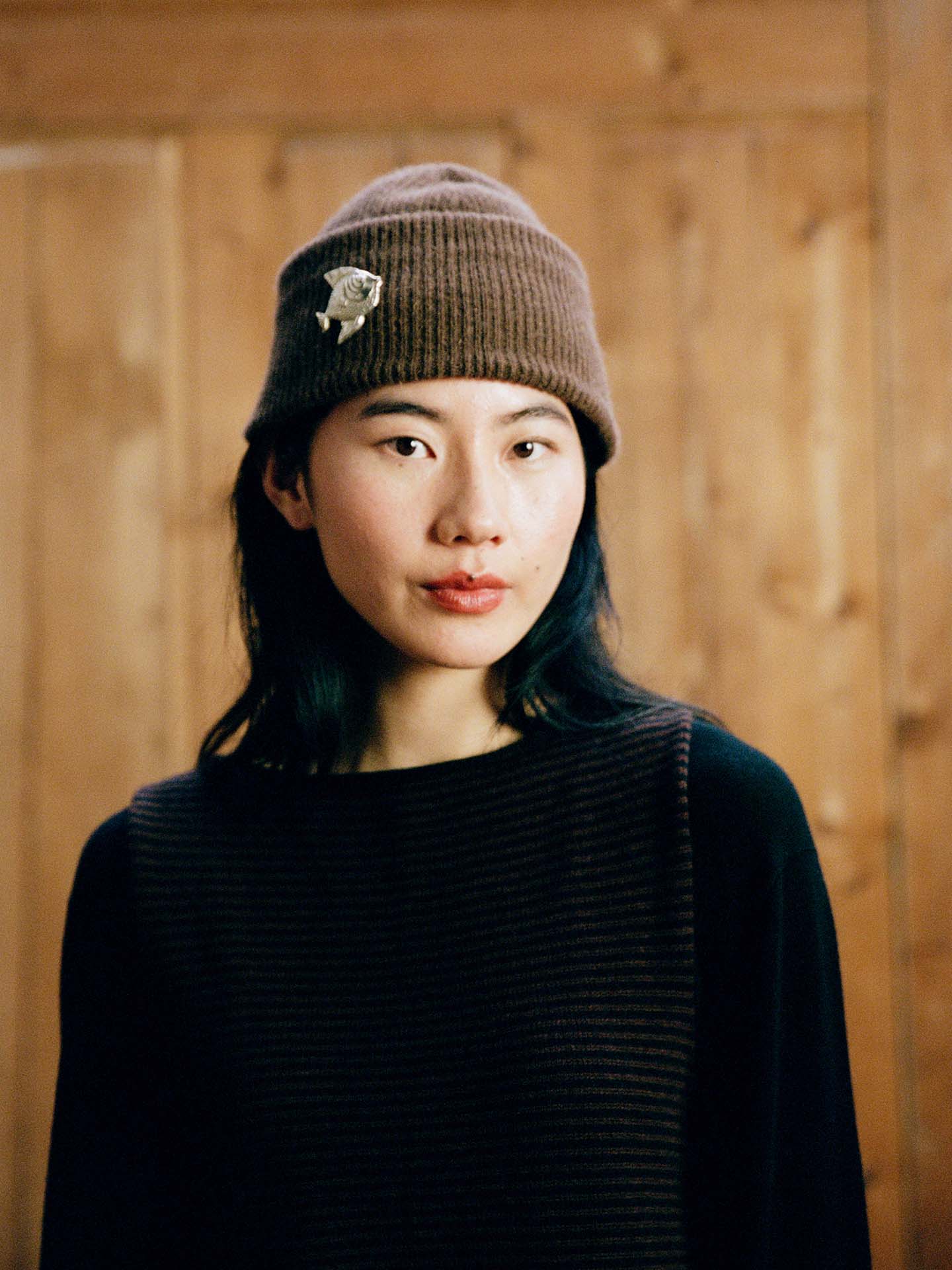 A person with shoulder-length dark hair wears the Hinoki Beanie – Chestnut, featuring a pin, and a dark striped top while standing in front of a wooden background.