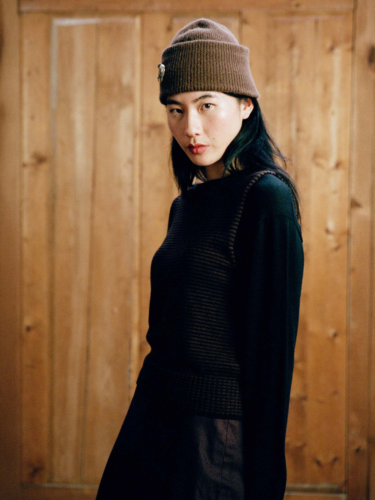 Person wearing the Hinoki Beanie in Chestnut and a dark sweater stands before a wooden wall, looking at the camera with a neutral expression.