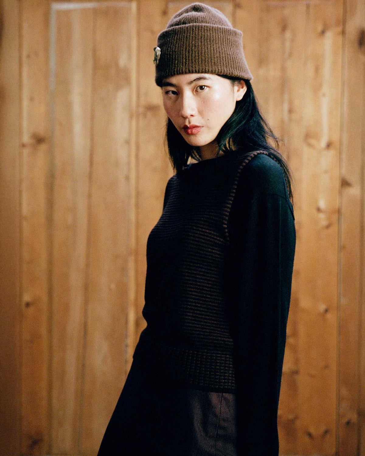 Person wearing the Hinoki Beanie in Chestnut and a dark sweater stands before a wooden wall, looking at the camera with a neutral expression.