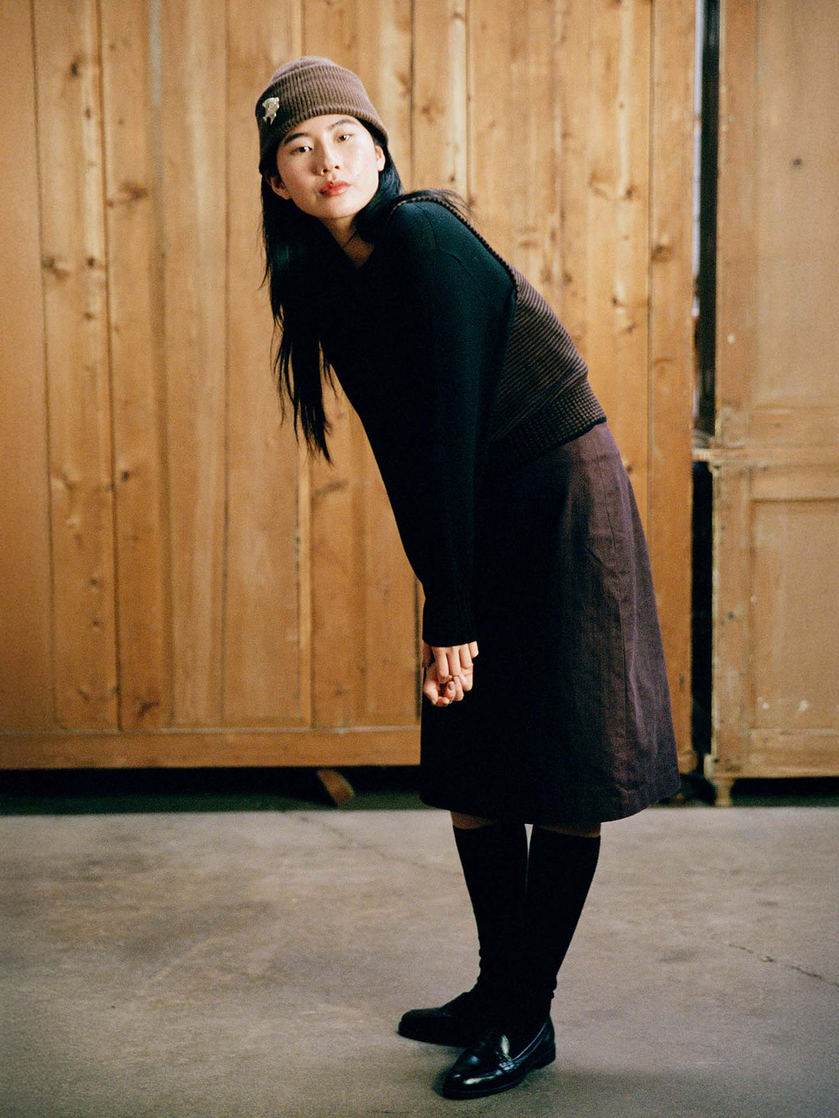A woman wears the Hinoki Beanie – Chestnut, paired with a brown skirt and black sweater, leaning forward slightly indoors on a concrete floor with wooden doors behind her.