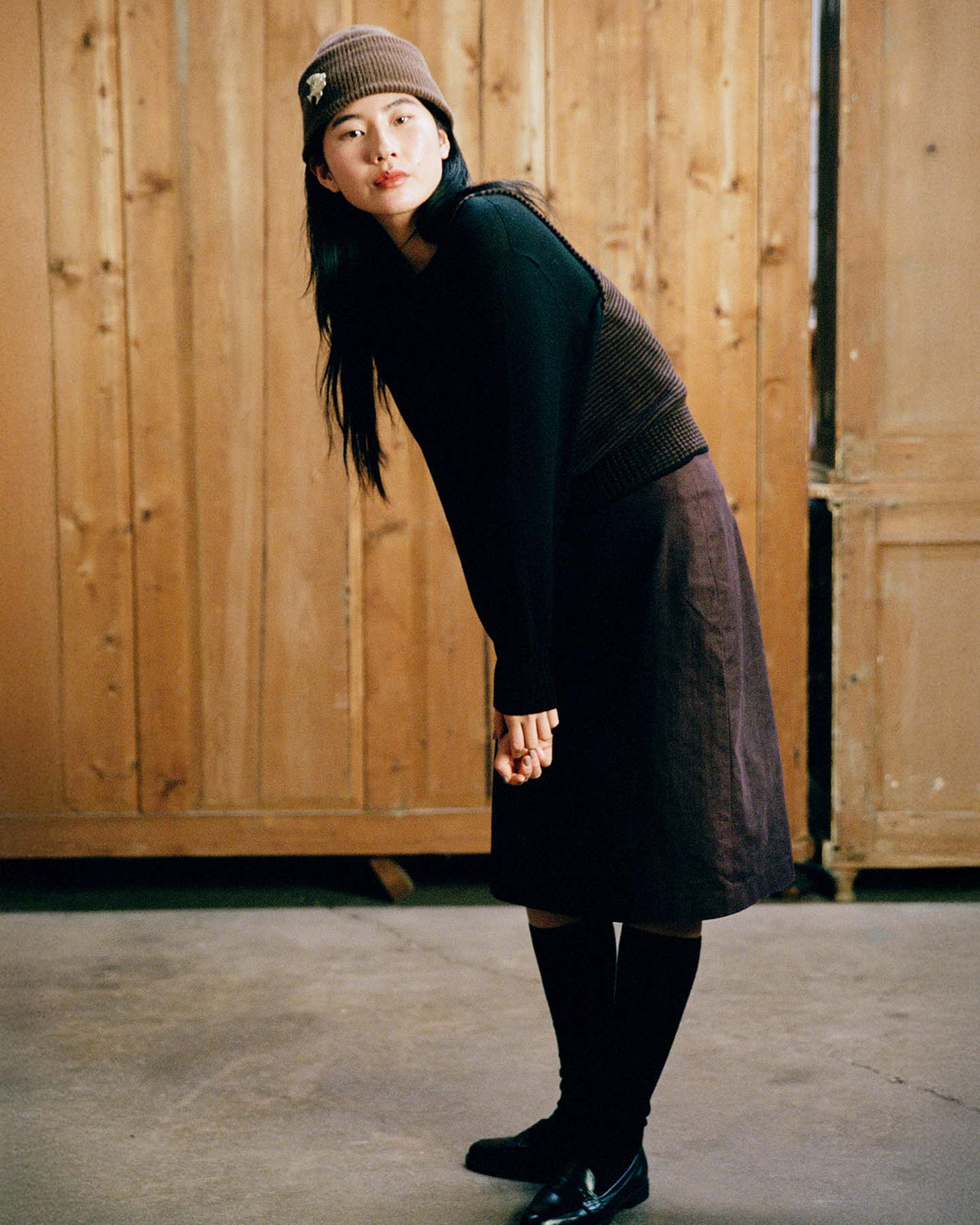 A woman wears the Hinoki Beanie – Chestnut, paired with a brown skirt and black sweater, leaning forward slightly indoors on a concrete floor with wooden doors behind her.