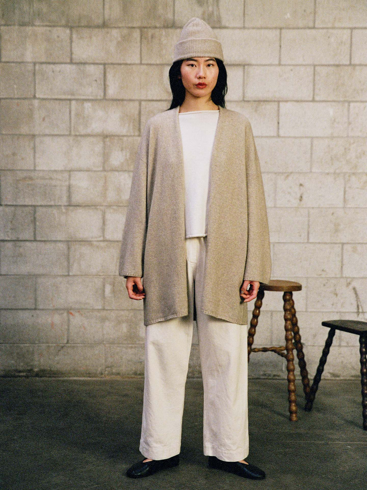 Indoors by a concrete block wall, a person wears the Hinoki Beanie – Natural, paired with a long beige cardigan, white top, white pants, and black shoes. Two wooden stools are in the background.