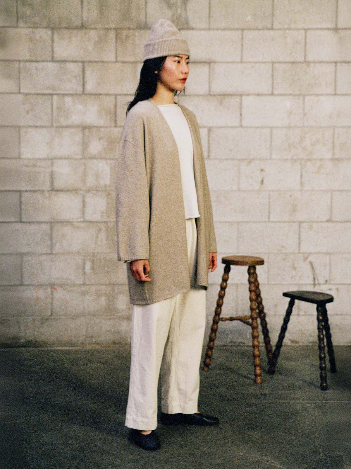 A person stands indoors by a cinderblock wall, wearing the Hinoki Beanie – Natural, a loose beige cardigan, white shirt, and white pants. Two wooden stools are visible in the background.