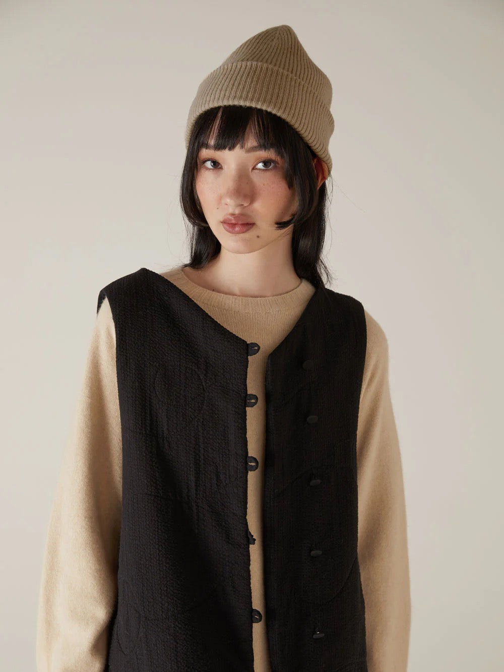 A person with long hair sports the Francie Hinoki Beanie in Natural, along with a beige sweater and black vest, standing against a neutral background.