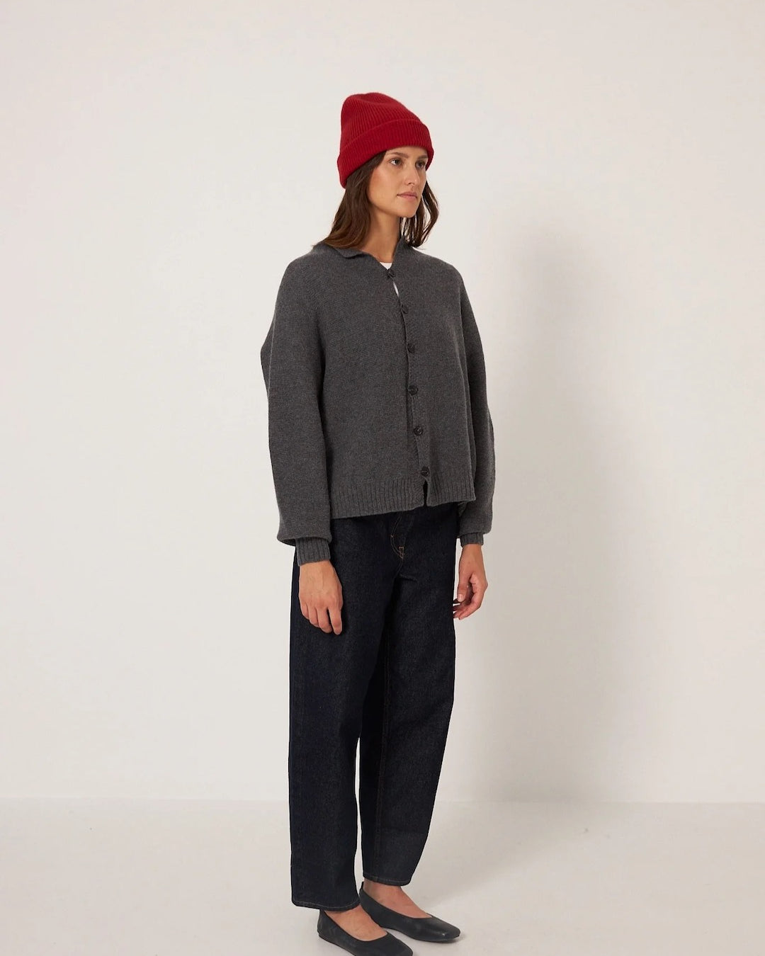 A person in a gray cardigan, black pants, and black flats stands against a plain background, wearing the Francie Hinoki Beanie in Poppy.