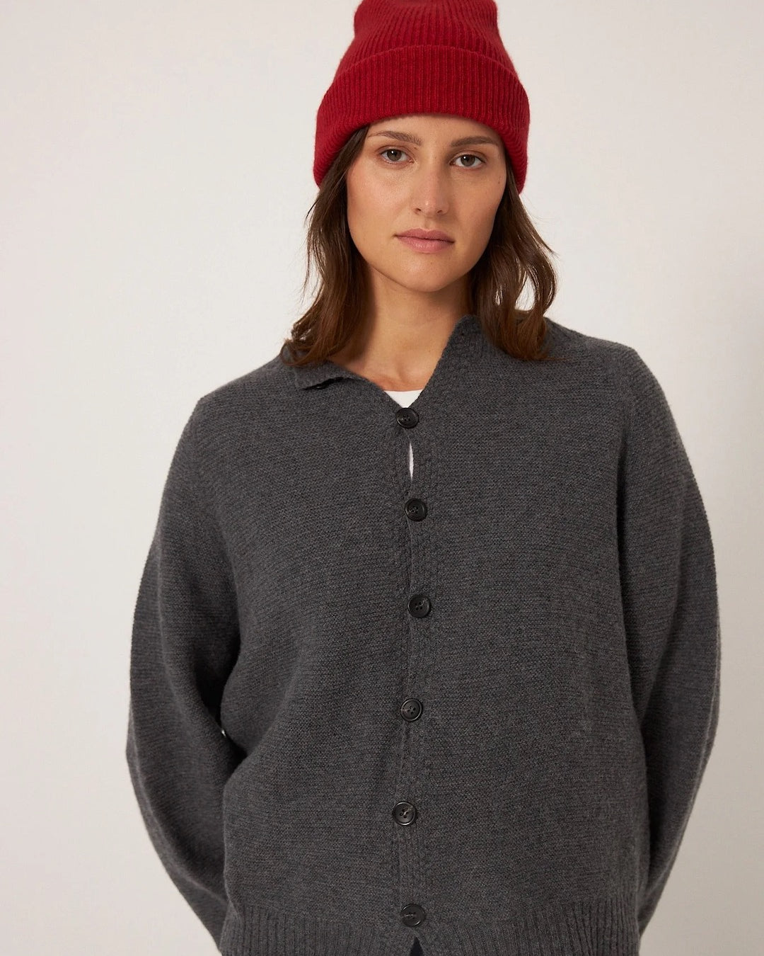 A person wears a Francie Hinoki Beanie in Poppy, a dark gray buttoned cardigan, and a white shirt, set against a plain background.